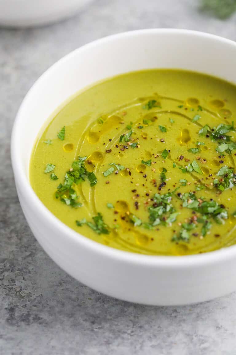 A White Bowl Filled With Creamy Pea And Asparagus Soup, Garnished With Chopped Herbs, Cracked Black Pepper, And A Drizzle Of Olive Oil, Sits On A Light Gray Surface.