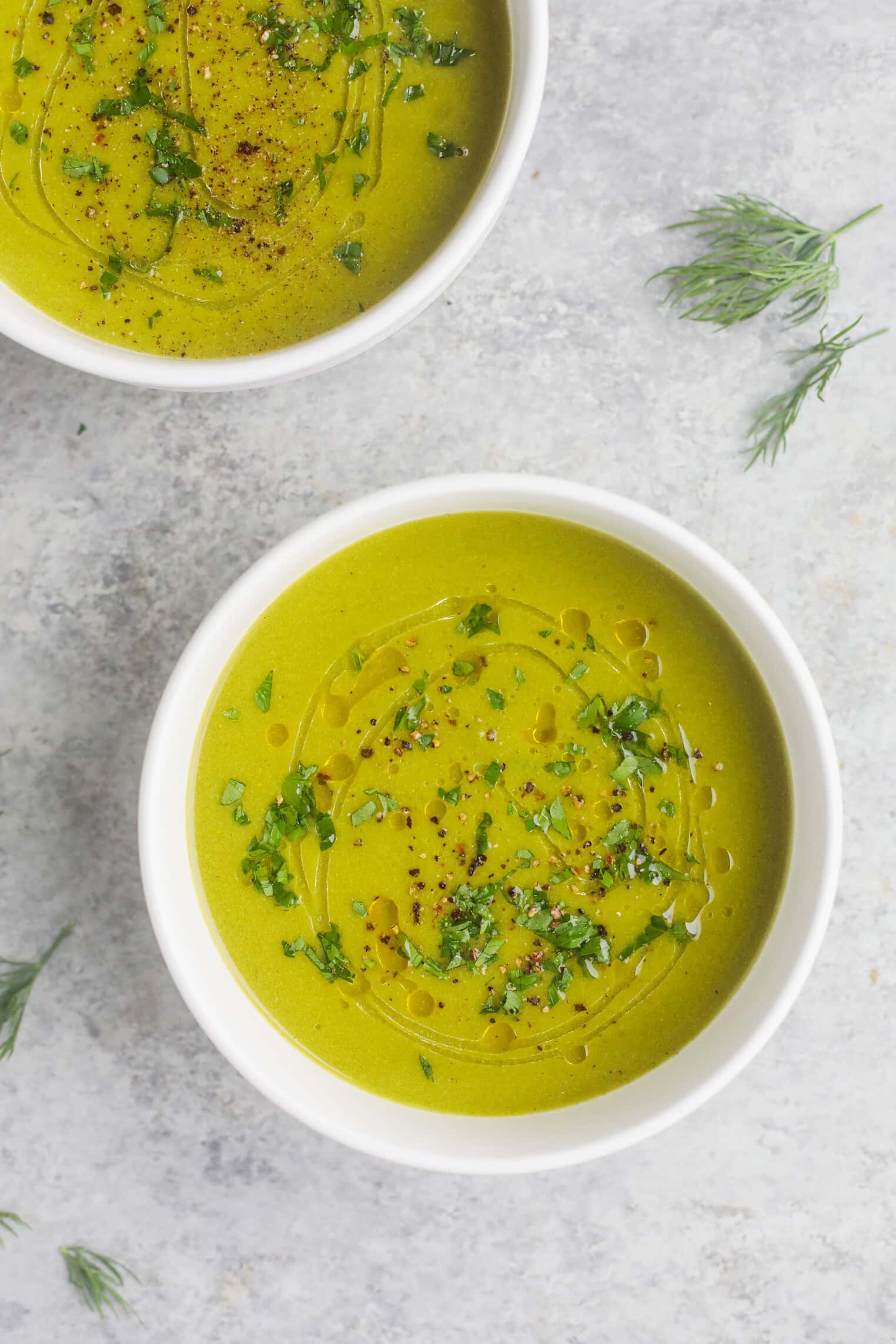 Two White Bowls Filled With Vibrant Pea And Asparagus Soup, Garnished With Chopped Herbs, Black Pepper, And A Drizzle Of Oil, Are Placed On A Light Gray Surface With Sprigs Of Fresh Dill Scattered Nearby.
