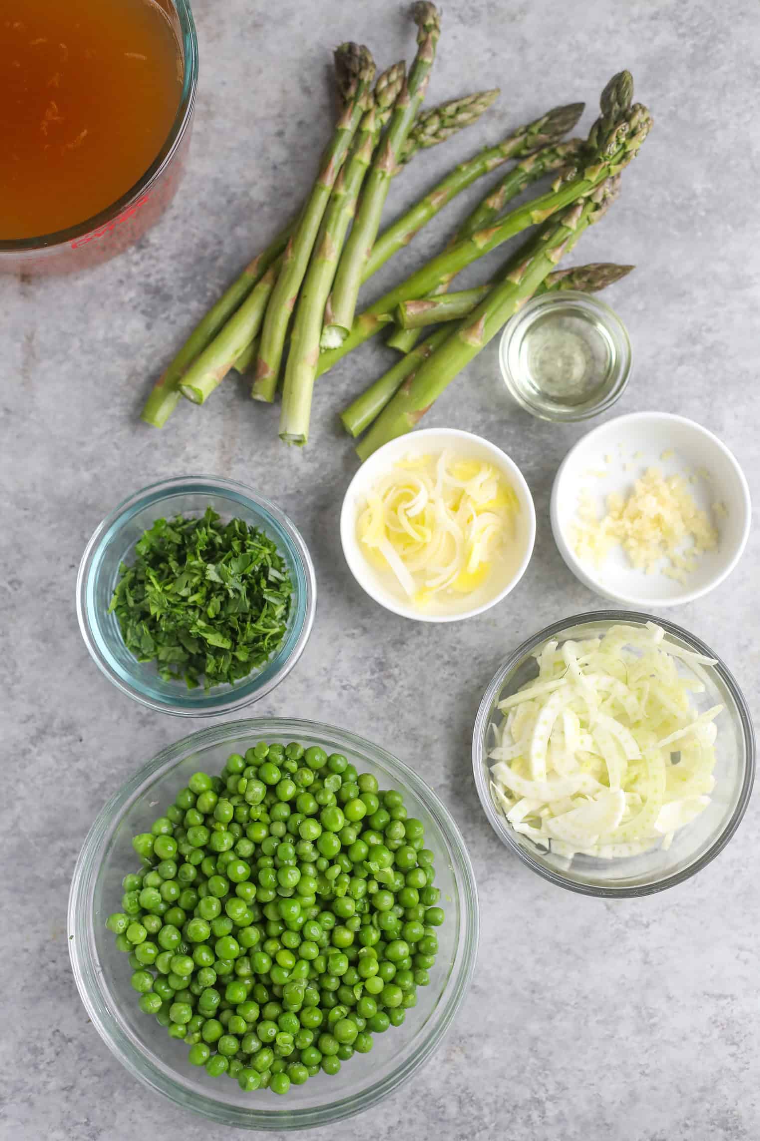 An Overhead View Of Raw Ingredients For Pea And Asparagus Soup On A Gray Surface: Asparagus Spears, A Bowl Of Green Peas, Sliced Fennel, Chopped Parsley, Grated Cheese, Minced Garlic, Oil, And A Cup Of Broth.