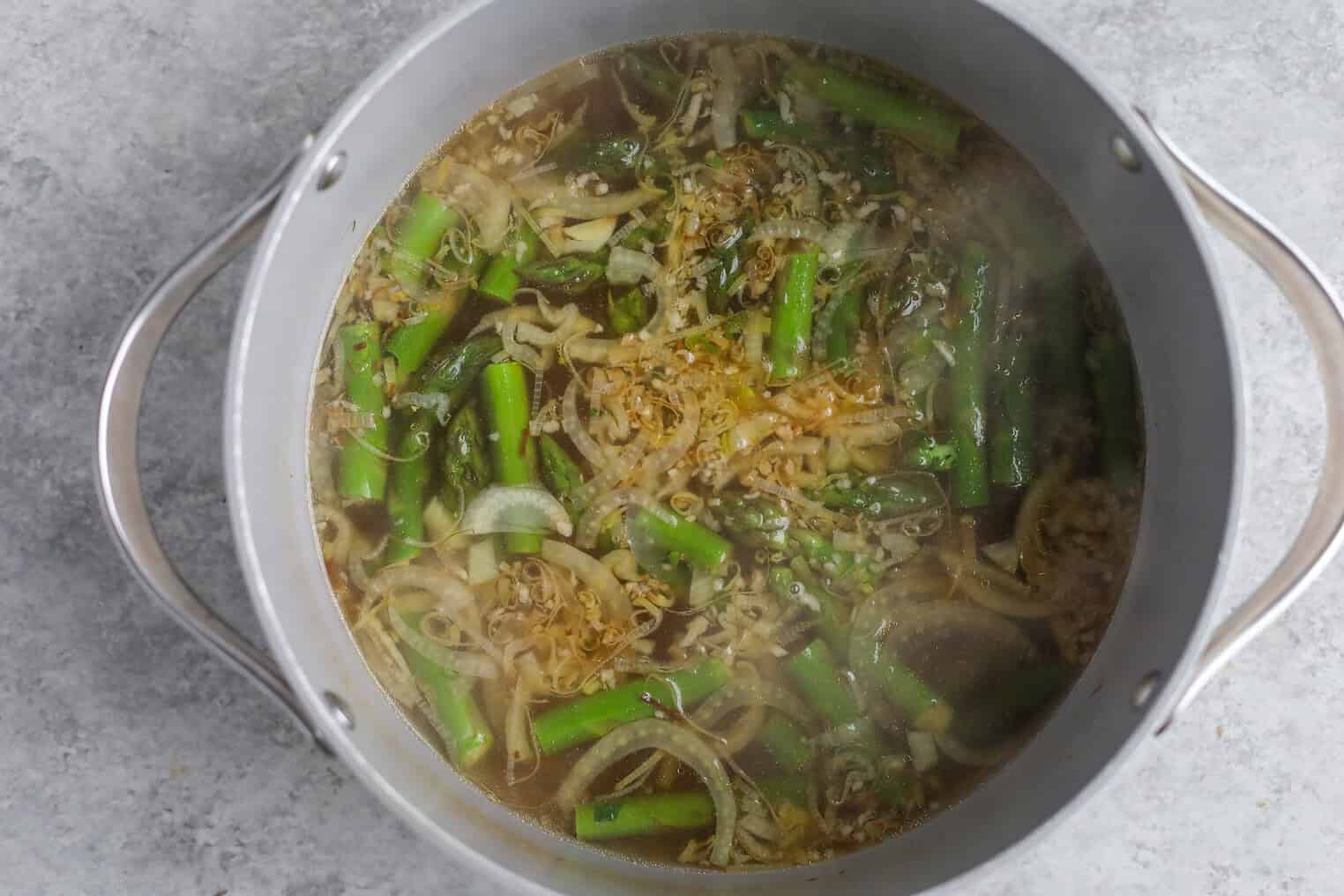 A Pot Of Pea And Asparagus Soup With Sliced Onions And Chopped Green Beans Simmers In Broth, Viewed From Above On A Light Gray Surface. Steam Is Rising From The Pot.