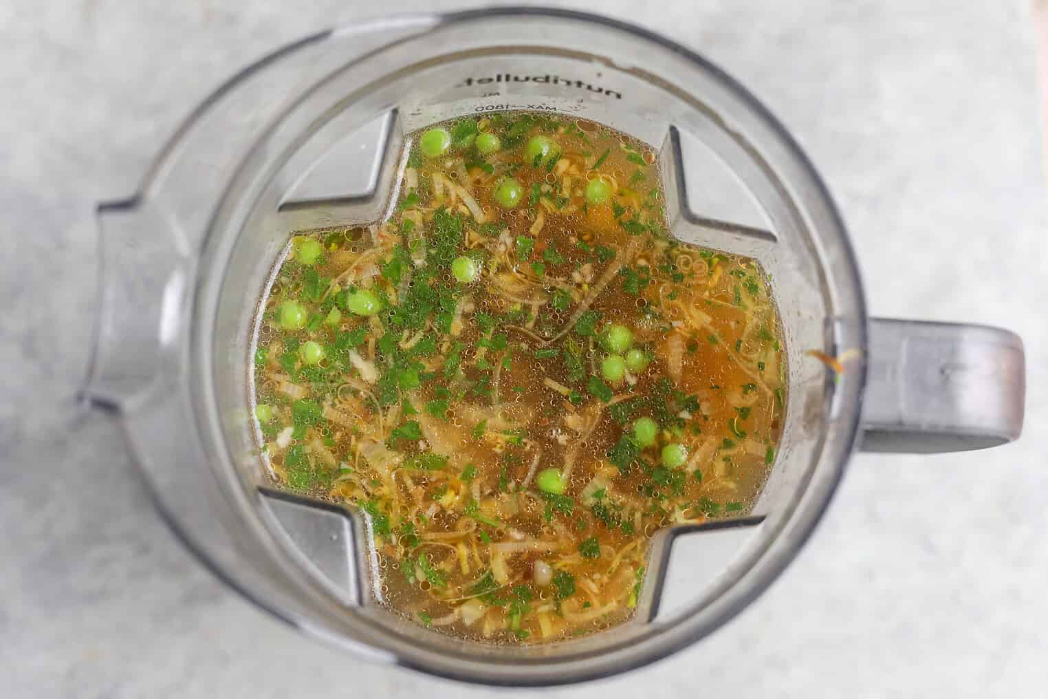Overhead View Of A Blender Containing Broth, Chopped Green Herbs, Peas, Asparagus, And Shredded Vegetables For A Fresh Pea And Asparagus Soup. The Blender Sits On A Light-Colored Surface, Ready To Blend Vibrant Flavors Together.