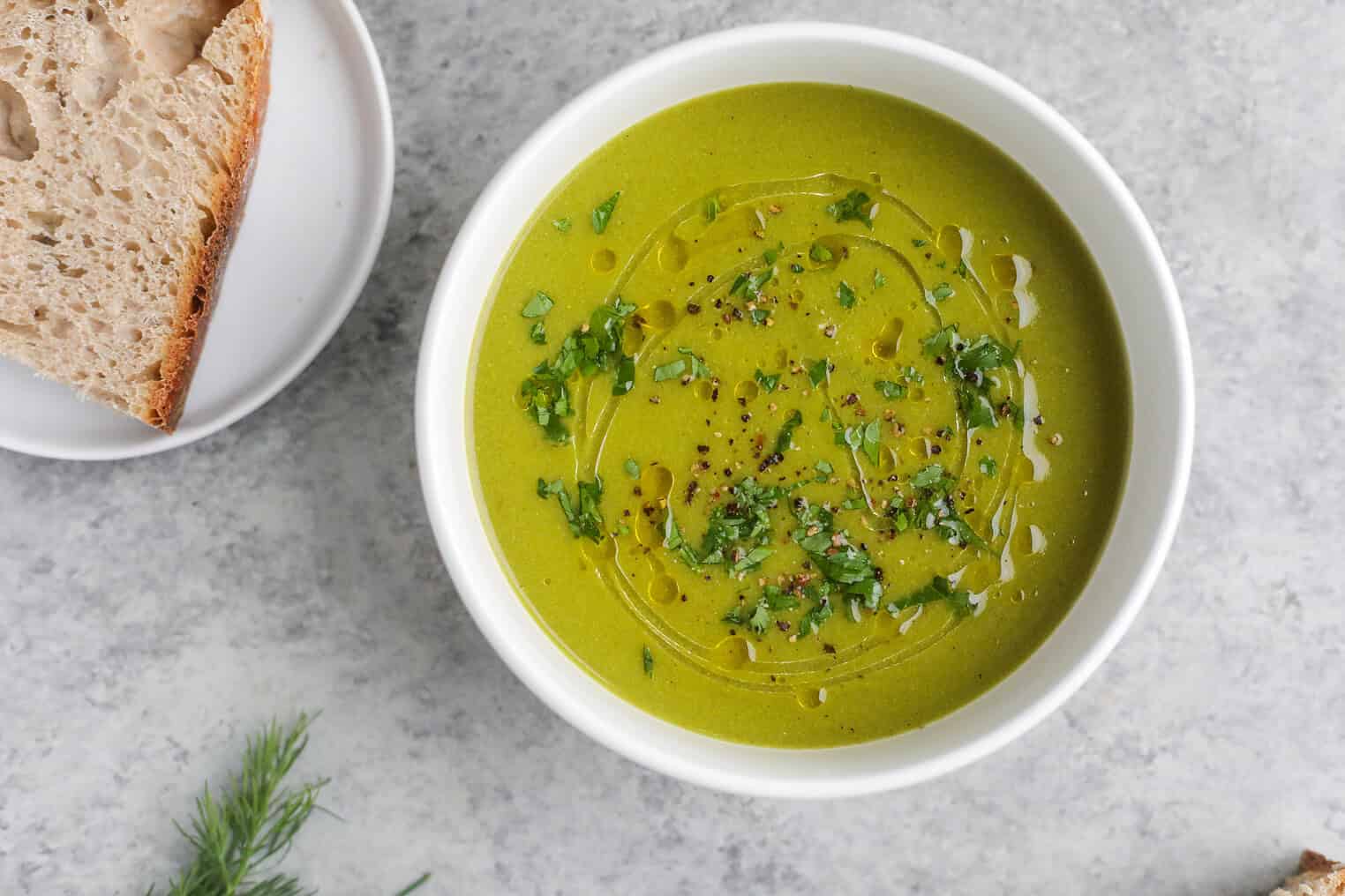 A Bowl Of Vibrant Pea And Asparagus Soup Garnished With Chopped Herbs, A Drizzle Of Olive Oil, And Cracked Pepper, Served On A Gray Surface With A Plate Of Rustic Bread On The Side.
