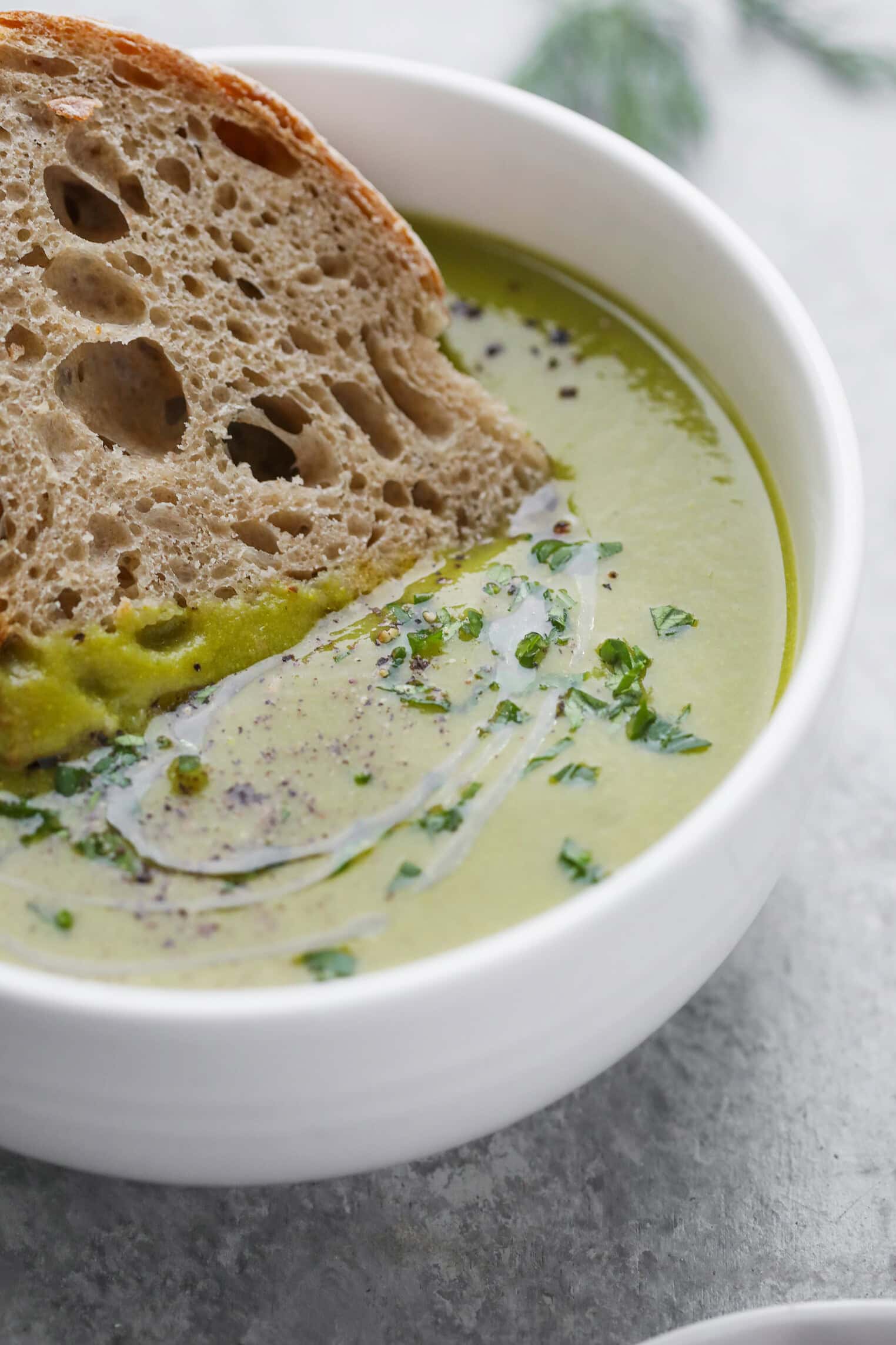 A Bowl Of Pea And Asparagus Soup Garnished With Herbs And Black Pepper, Topped With A Swirl Of Cream, And Served With A Slice Of Rustic Bread.