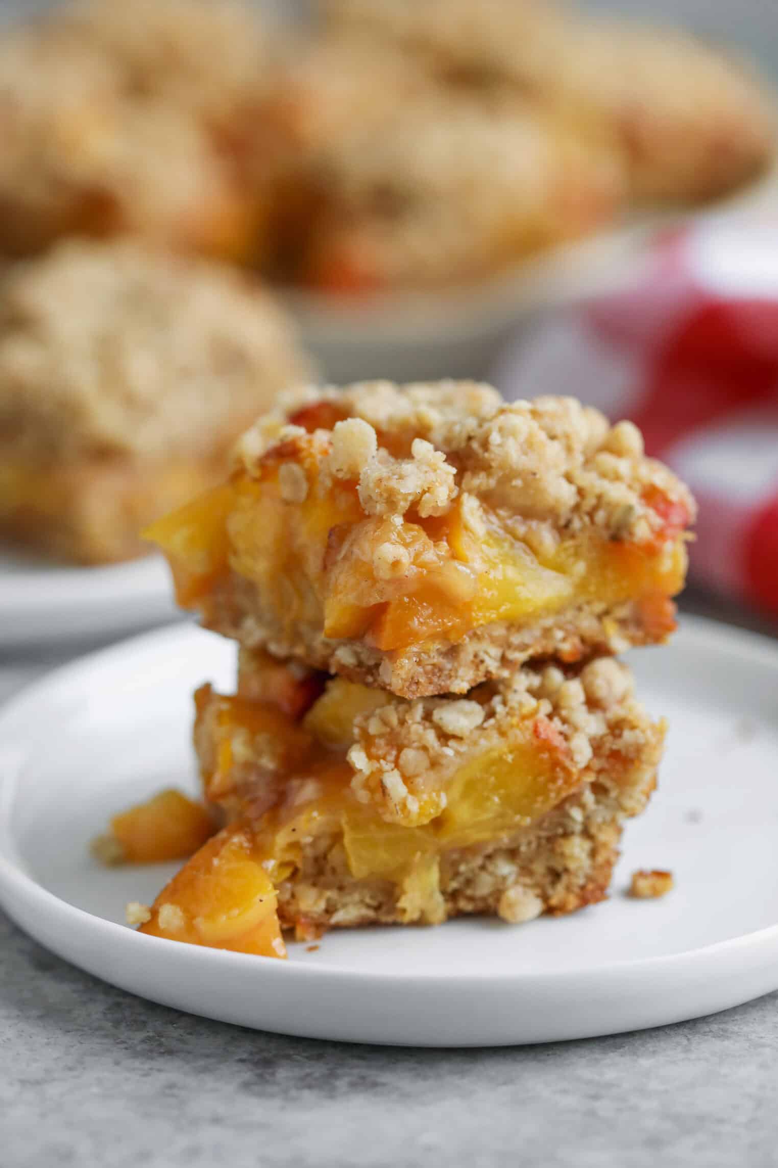 Peach Pie Bars 1 Two Peach Pie Bars Stacked On A White Plate, Displaying Layers Of Golden Peach Filling And Crumbly Topping, With Blurred Bars And Red Fruit In The Background.