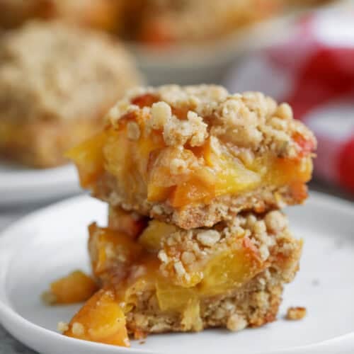 Peach Pie Bars 13 Two Peach Pie Bars Stacked On A White Plate, Displaying Layers Of Golden Peach Filling And Crumbly Topping, With Blurred Bars And Red Fruit In The Background.