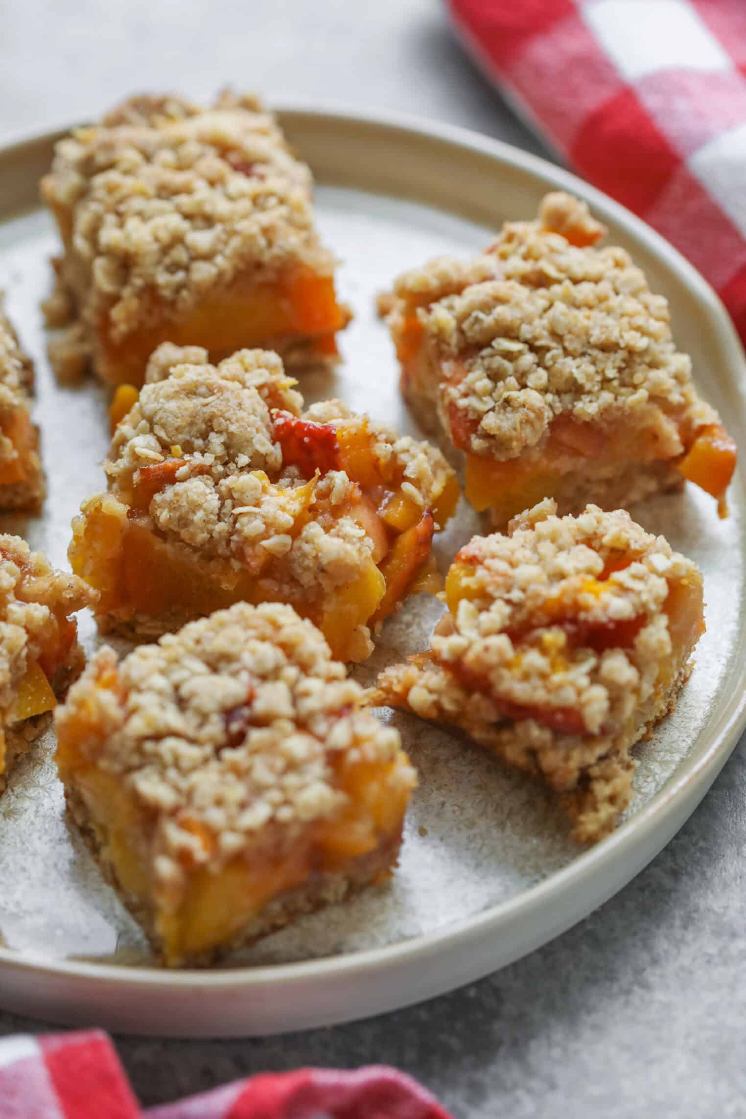 Peach Pie Bars 2 Six Peach Pie Bars With A Golden Oat Topping Are Arranged On A Round Beige Plate. The Dessert Bars Appear Moist And Crumbly, And A Red-And-White Checkered Cloth Is Partially Visible Beside The Plate.