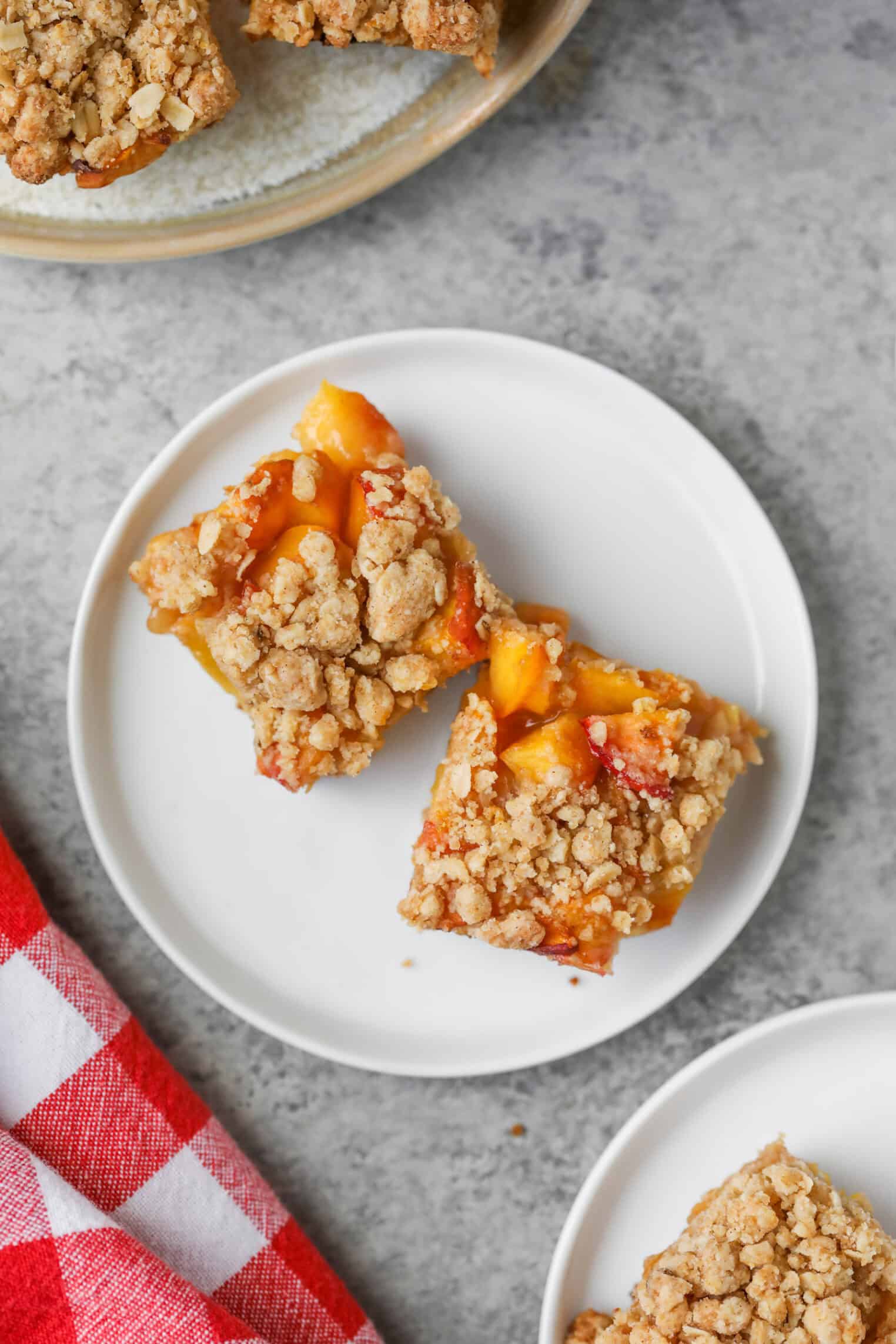 Peach Pie Bars 3 Two Peach Pie Bars Sit On A White Plate, Featuring A Golden Oat Crumb Topping And Visible Pieces Of Peach. A Red And White Checkered Cloth And Part Of Another Plate Are Nearby On A Gray Surface.
