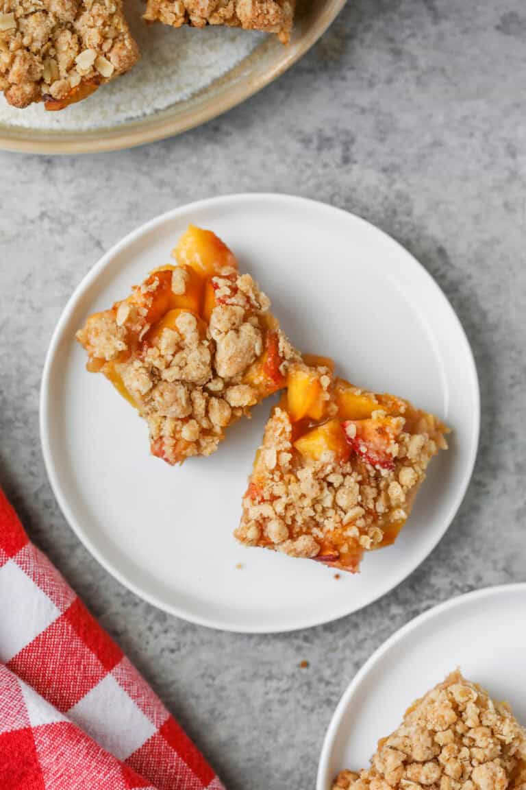 Two Peach Pie Bars Sit On A White Plate, Featuring A Golden Oat Crumb Topping And Visible Pieces Of Peach. A Red And White Checkered Cloth And Part Of Another Plate Are Nearby On A Gray Surface.