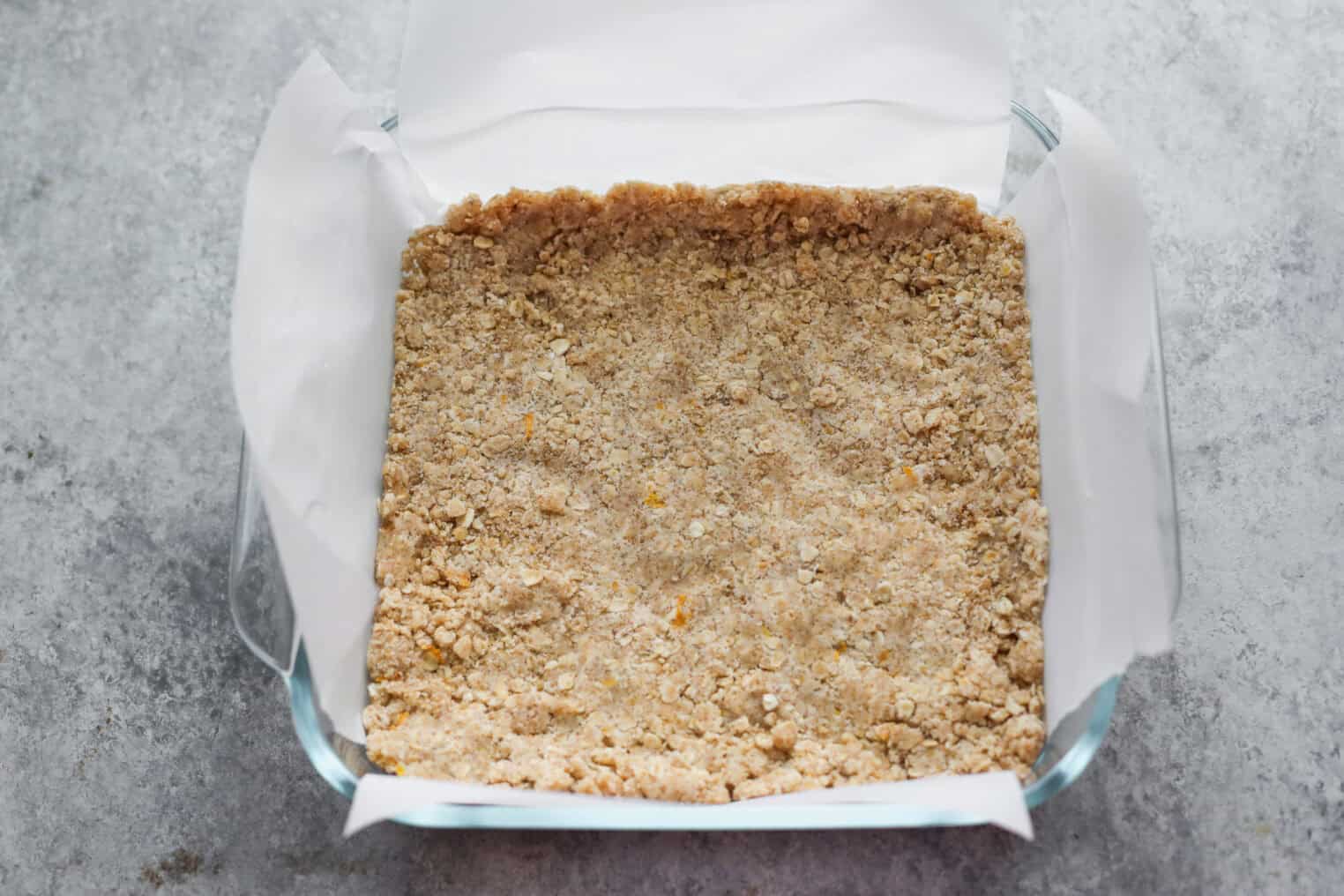 Peach Pie Bars 7 A Glass Baking Dish Lined With Parchment Paper Holds A Pressed, Crumbly Oat Mixture—The Perfect Base For Delicious Peach Pie Bars—Resting On A Gray Countertop.