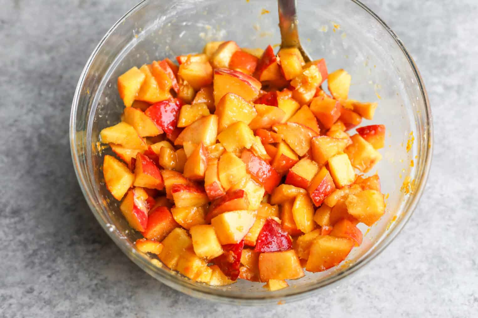 Peach Pie Bars 8 A Clear Glass Bowl Filled With Chopped Nectarines Mixed With A Yellow-Orange Sauce, Reminiscent Of The Filling In Peach Pie Bars, With A Spoon In The Bowl, Placed On A Gray Countertop.