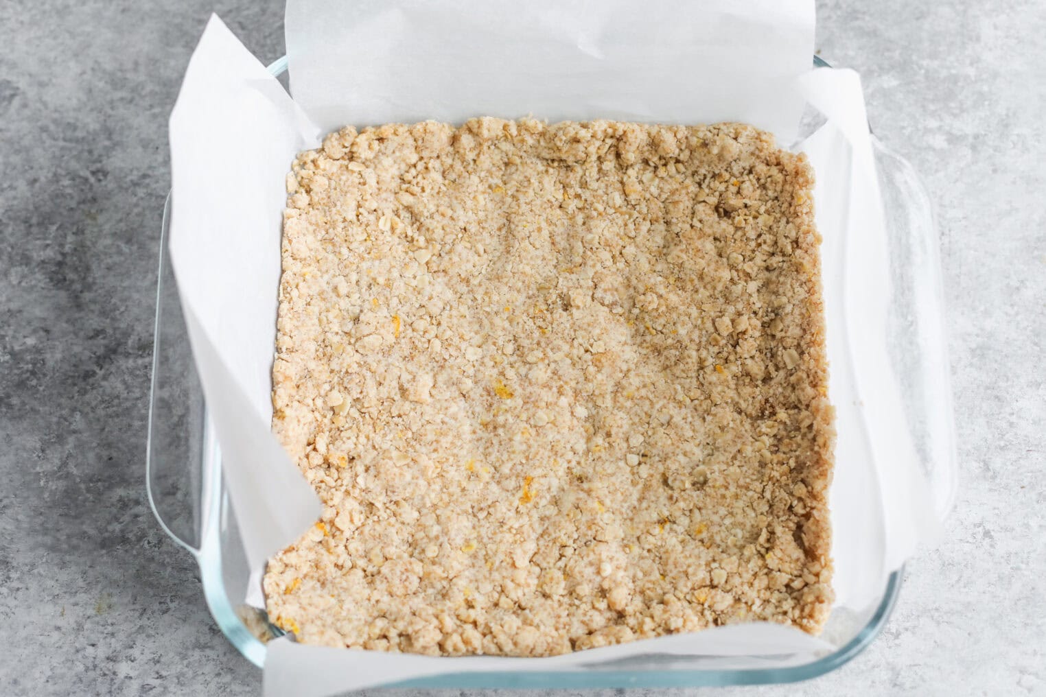 Peach Pie Bars 6 A Glass Baking Dish Lined With Parchment Paper Is Filled With An Even Layer Of Crumbly Oat Mixture For Peach Pie Bars, Set On A Gray Countertop, Ready To Be Baked.