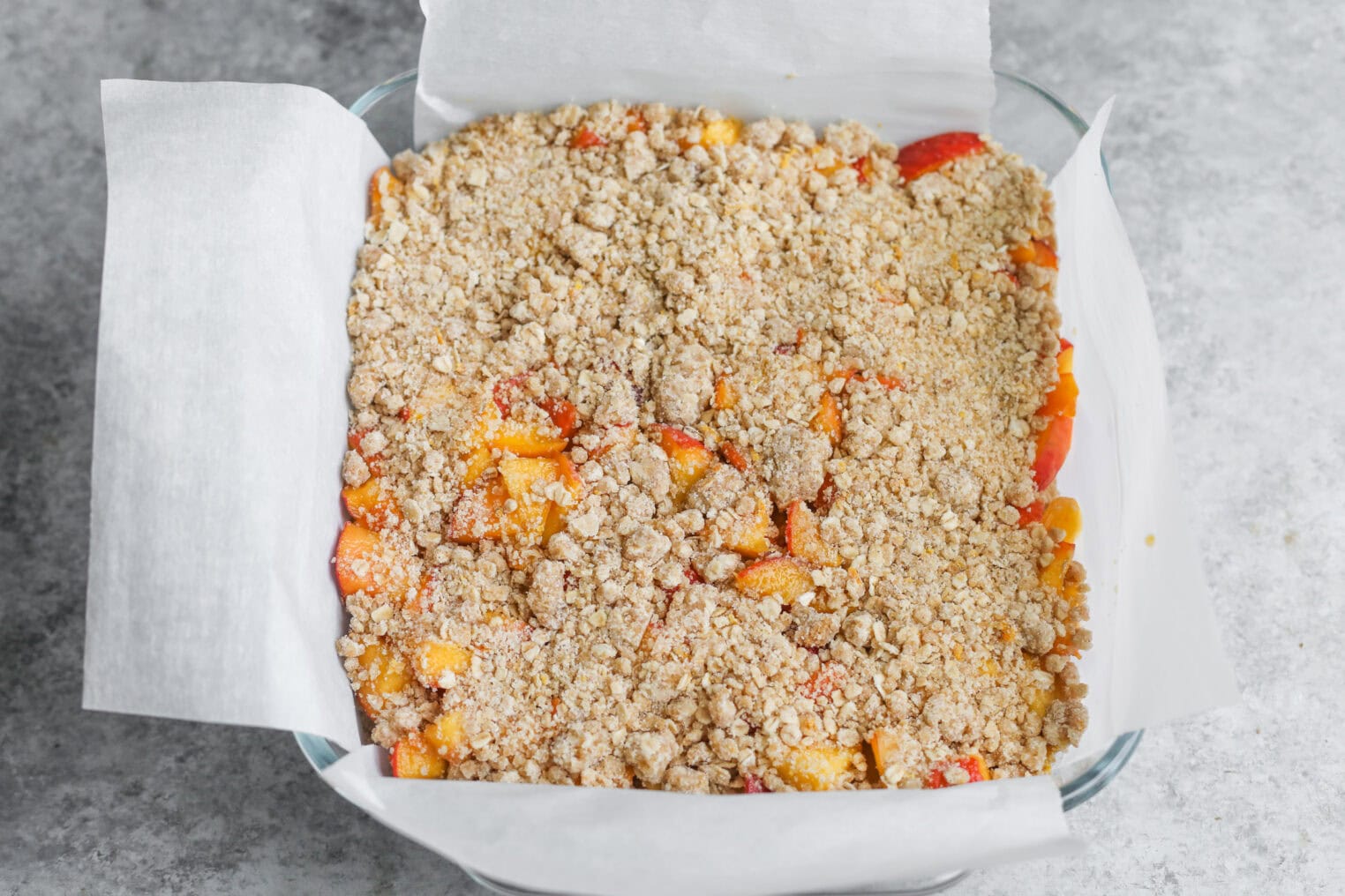 Peach Pie Bars 9 A Glass Baking Dish Lined With Parchment Paper Holds Unbaked Peach Pie Bars, Showing A Layer Of Chopped Fruit Beneath A Crumbly Oat And Sugar Topping On A Gray Countertop.