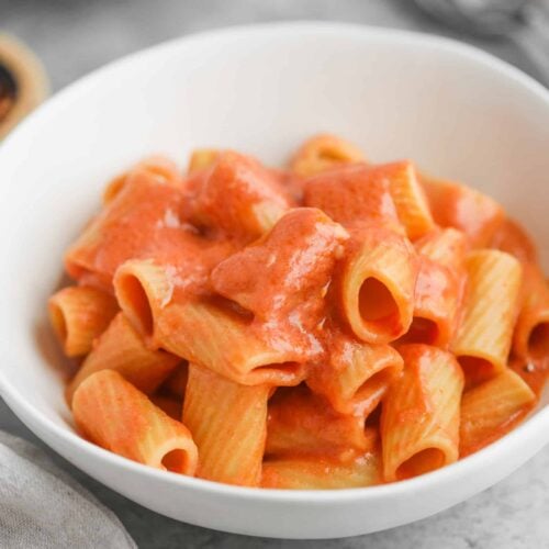 A White Bowl Filled With Rigatoni Pasta Coated In A Velvety Pink Tomato Sauce Reminiscent Of Penne Alla Vodka Rests On A Gray Surface, Accompanied By A Fork And Napkin In The Background.