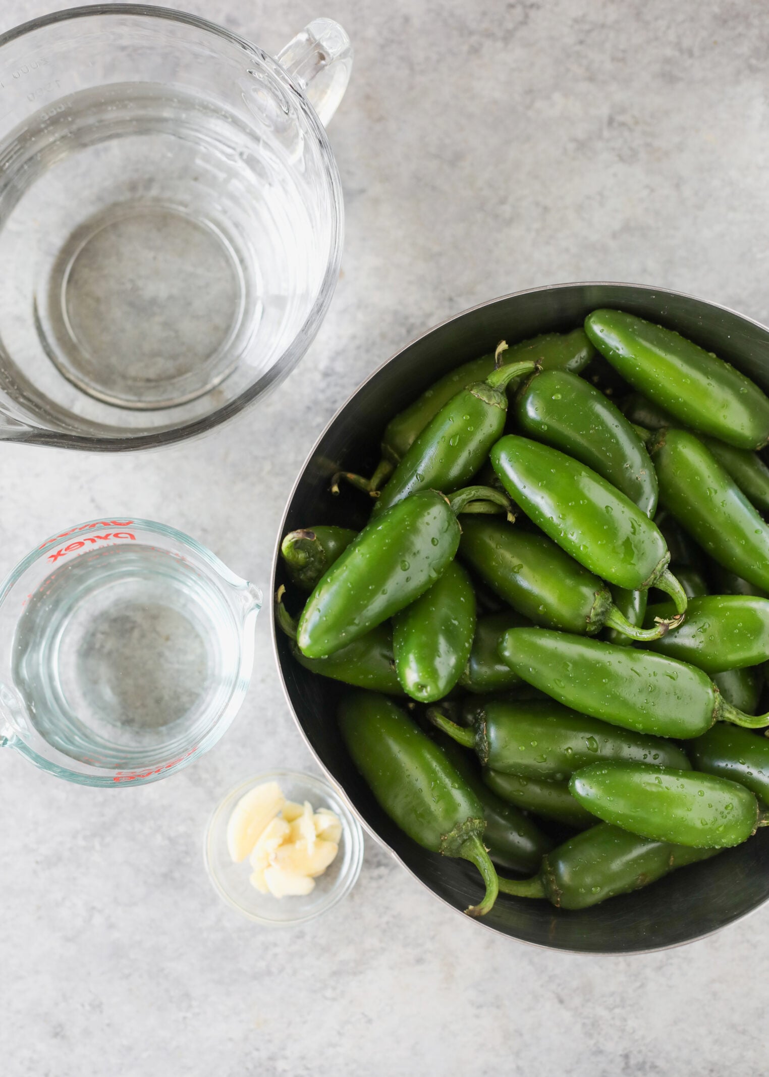 Pickled Jalapeños (Canning Recipe) 3 A Bowl Of Fresh Green Jalapeños, Ready To Be Transformed Into Tangy Pickled Jalapenos, Sits Beside A Small Glass Of Garlic Cloves, A Large Measuring Cup Of Water, And A Smaller Measuring Cup Of Clear Liquid On A Gray Countertop.