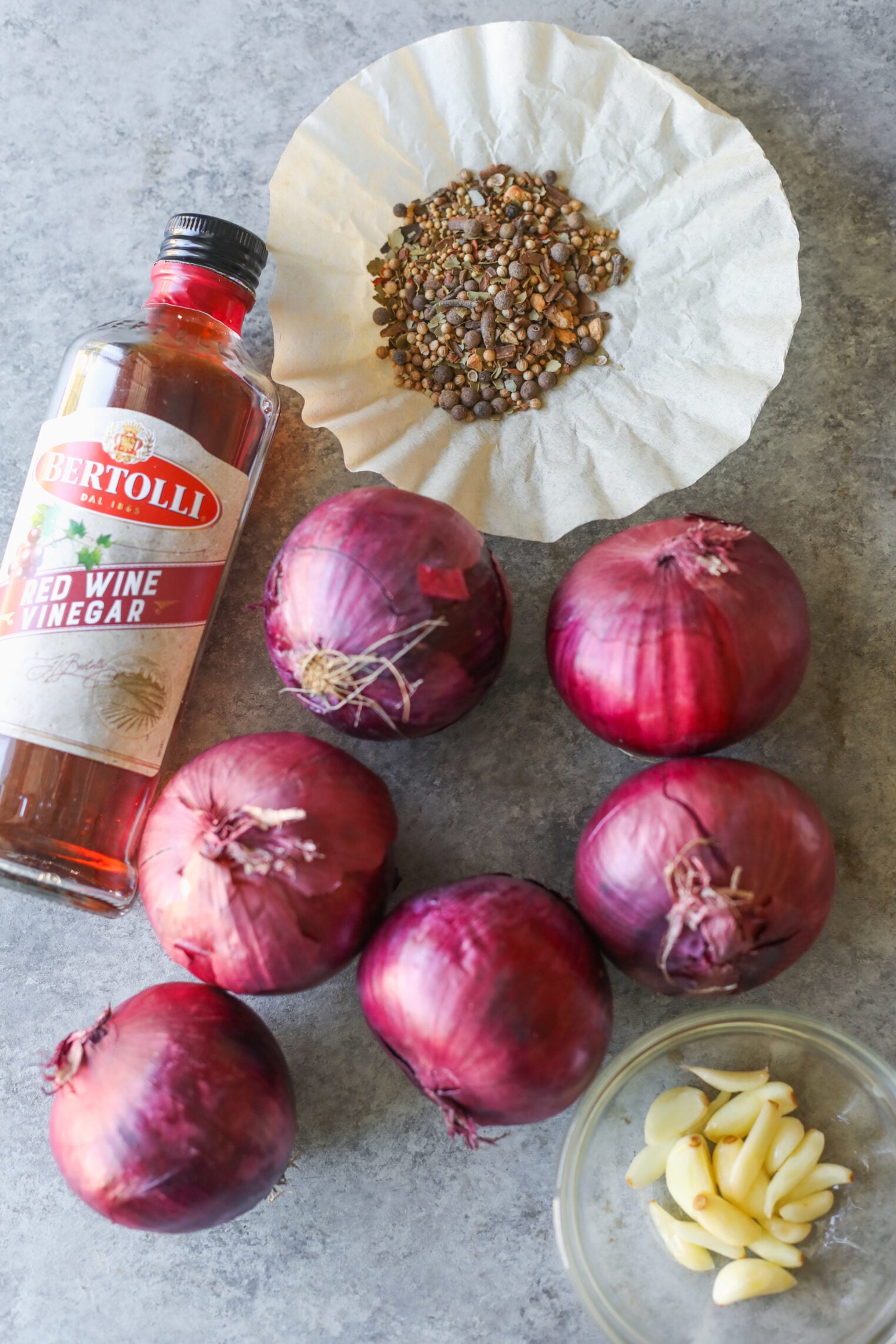 Pickled Red Onions 3 A Bottle Of Bertolli Red Wine Vinegar, Five Red Onions, A Small Bowl Of Garlic Cloves, And A Coffee Filter Holding Pickling Spices To Make Pickled Red Onions.