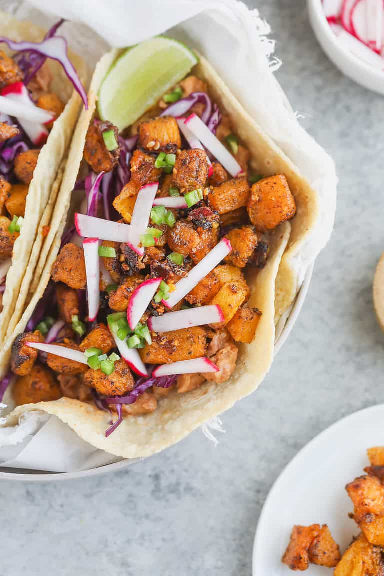 A Close-Up Of A Pinto Bean Taco Filled With Spiced Roasted Sweet Potatoes, Shredded Purple Cabbage, Sliced Radishes, Chopped Green Onions, And A Lime Wedge In A Soft Tortilla, Resting On A White Cloth.