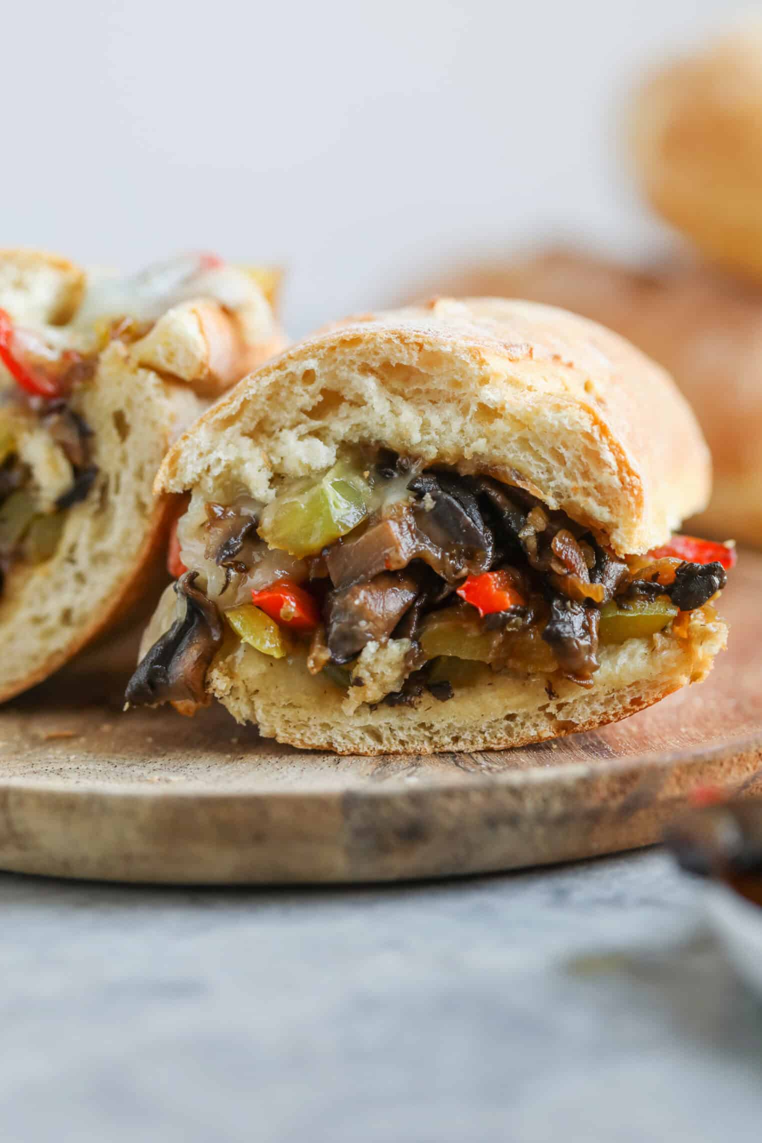 Portobello Cheesesteak Sandwiches 1 A Close-Up Of A Portobello Cheesesteak Sandwich With Sautéed Mushrooms, Red Peppers, And Green Vegetables On A Sliced Baguette, Placed On A Wooden Serving Board.