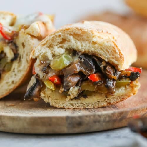 Portobello Cheesesteak Sandwiches 11 A Close-Up Of A Portobello Cheesesteak Sandwich With Sautéed Mushrooms, Red Peppers, And Green Vegetables On A Sliced Baguette, Placed On A Wooden Serving Board.