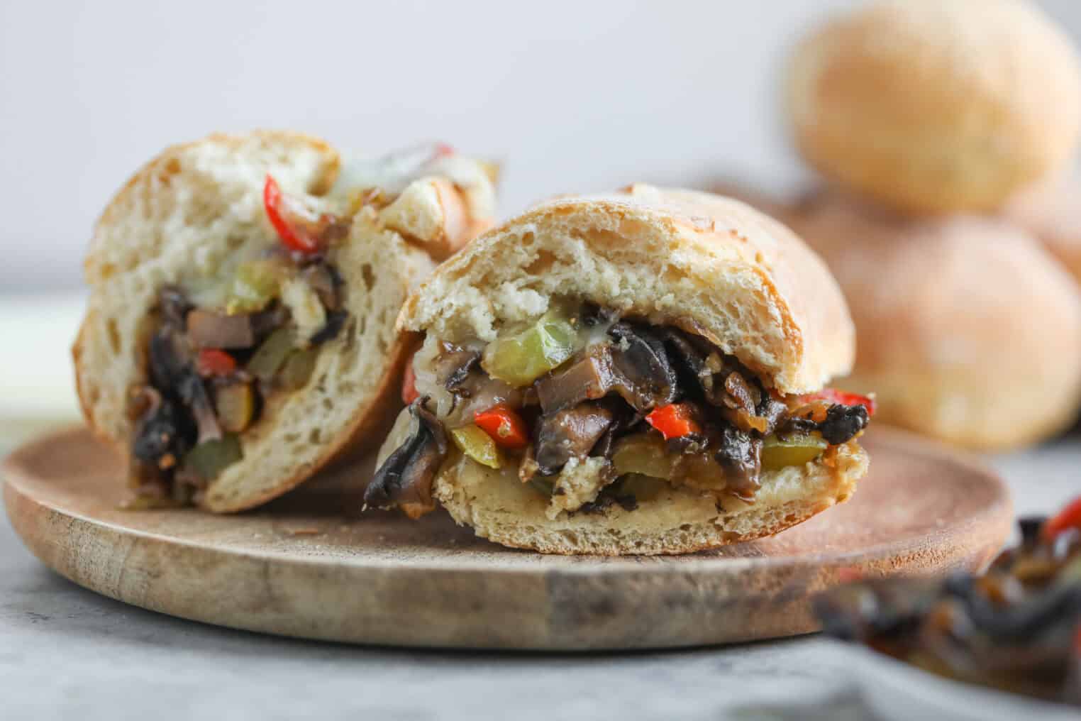 Portobello Cheesesteak Sandwiches 8 A Close-Up Of Portobello Cheesesteak Sandwiches Cut In Half On A Wooden Plate, Filled With Sautéed Vegetables Like Mushrooms, Onions, And Red Peppers. The Bread Looks Crusty And Fresh. Blurred Rolls Are Visible In The Background.