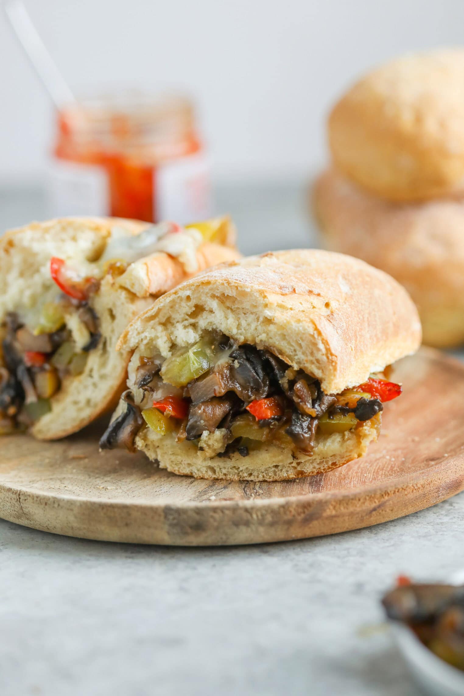 Portobello Cheesesteak Sandwiches 9 A Ciabatta Sandwich Inspired By Portobello Cheesesteak Sandwiches, Filled With Sautéed Mushrooms, Bell Peppers, And Onions, Is Cut In Half On A Wooden Board. A Jar Of Condiment And Bread Rolls Are In The Background.