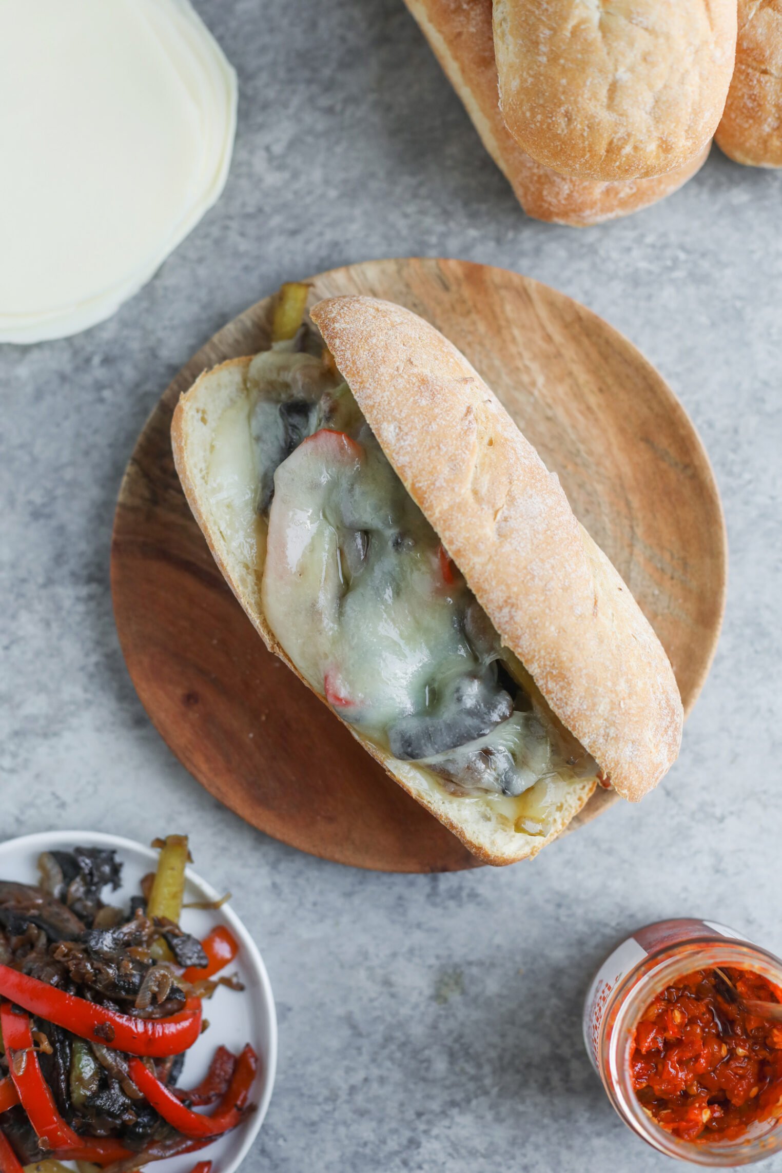 Portobello Cheesesteak Sandwiches 2 A Portobello Cheesesteak Sandwich Sits On A Wooden Plate, Filled With Melted Cheese, Sautéed Peppers, Onions, And Mushrooms. Nearby Are A Stack Of Rolls, Cooked Veggies, A Jar Of Sauce, And Cheese Slices.