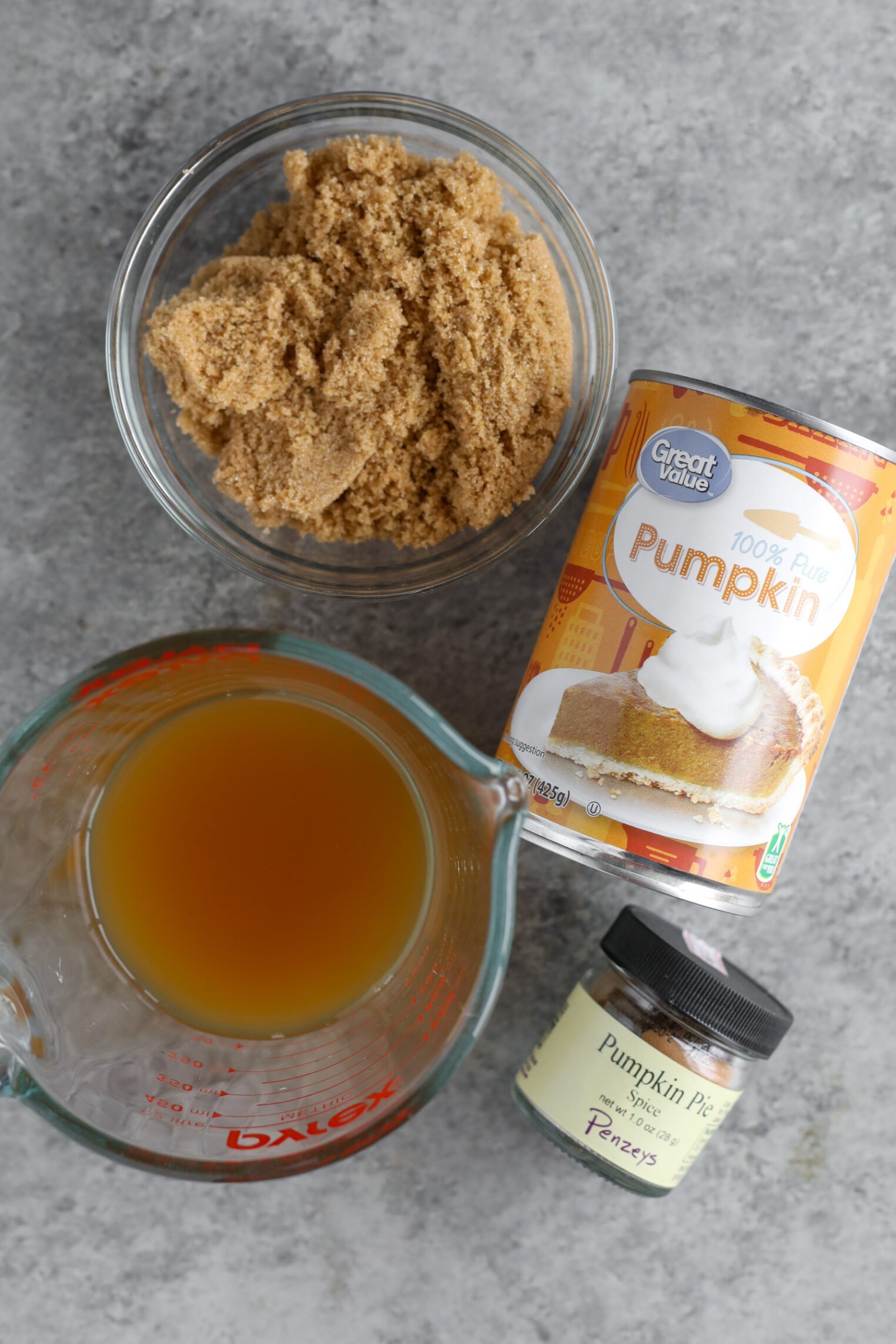 Pumpkin Butter 2 A Bowl Of Brown Sugar, A Can Of Pumpkin Puree, A Measuring Cup Of Apple Cider, And A Small Jar Of Pumpkin Pie Spice Are Arranged On A Gray Countertop.