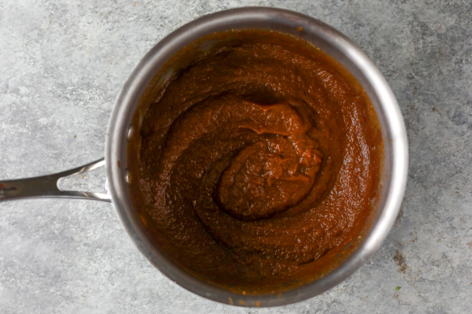 Pumpkin Butter 4 A Stainless Steel Saucepan Filled With Thick, Dark Red Tomato Sauce Sits On A Gray Textured Surface. The Sauce Appears Smooth And Well-Blended.