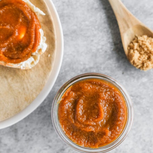 Pumpkin Butter 8 A Jar Of Pumpkin Butter On A Gray Surface, Next To A Slice Of Bread With Pumpkin Butter And Cream Cheese, A Wooden Spoon With Brown Sugar, And Cinnamon Sticks.