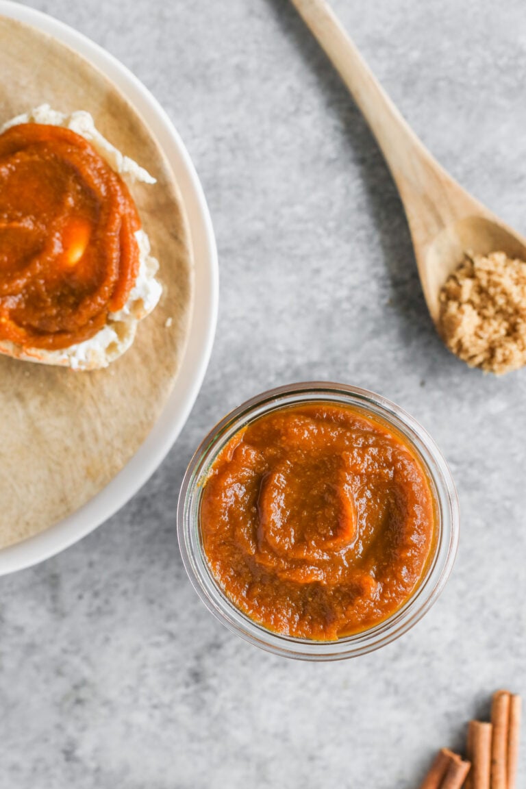 A Jar Of Pumpkin Butter On A Gray Surface, Next To A Slice Of Bread With Pumpkin Butter And Cream Cheese, A Wooden Spoon With Brown Sugar, And Cinnamon Sticks.