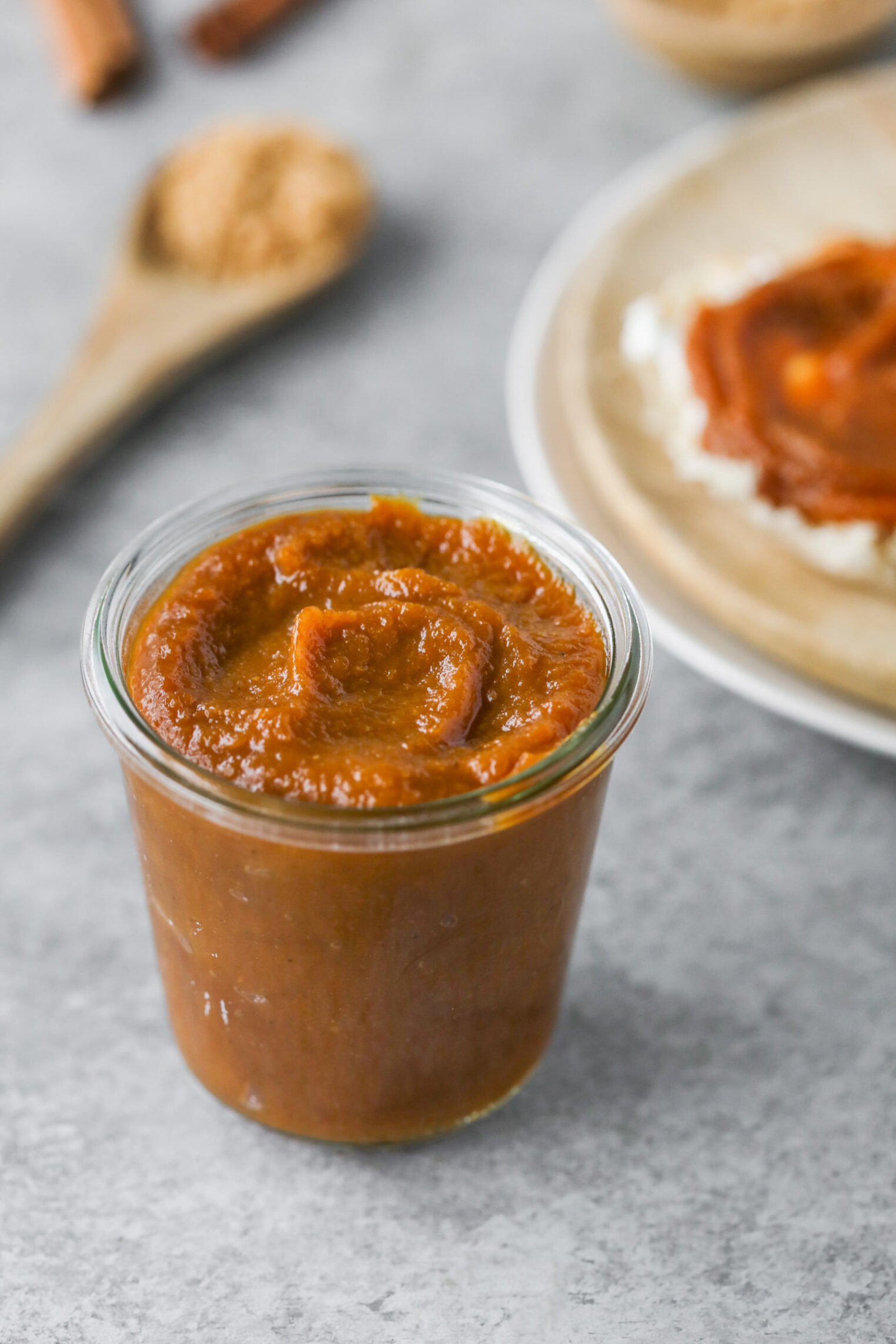Pumpkin Butter 1 A Small Glass Jar Filled With Thick, Brown Apple Butter Sits On A Gray Surface; In The Background Are A Wooden Spoon With Brown Sugar And A Plate With Apple Butter Spread On Toast.