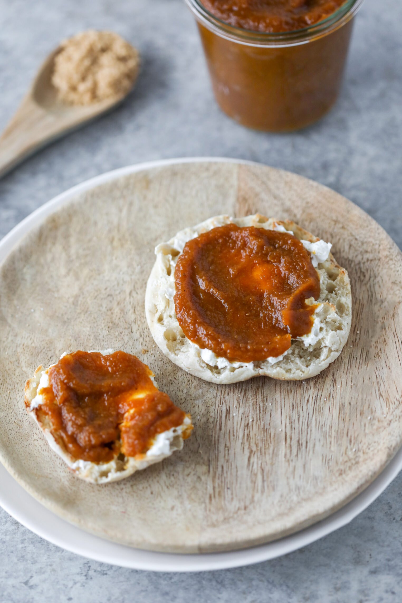 Pumpkin Butter 5 A Wooden Plate Holds An English Muffin Split In Half, Topped With Creamy Cheese And A Spread Of Apple Butter. In The Background, There’s A Jar Of Apple Butter And A Wooden Spoon With Brown Sugar.