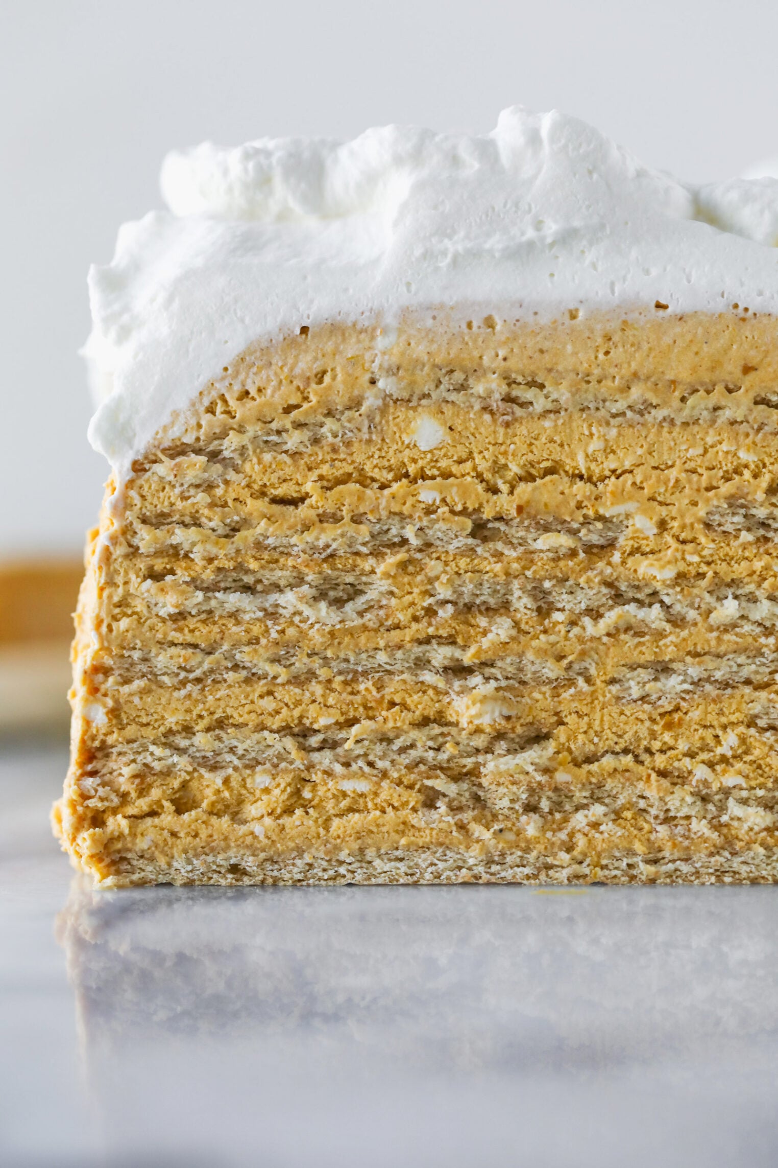 A Close-Up Of A Layered Dessert, Likely An Icebox Cake, Featuring Multiple Thin Cracker Or Cookie Layers Filled With A Creamy Orange-Tinted Mixture And Topped With A Thick Layer Of Whipped Cream.