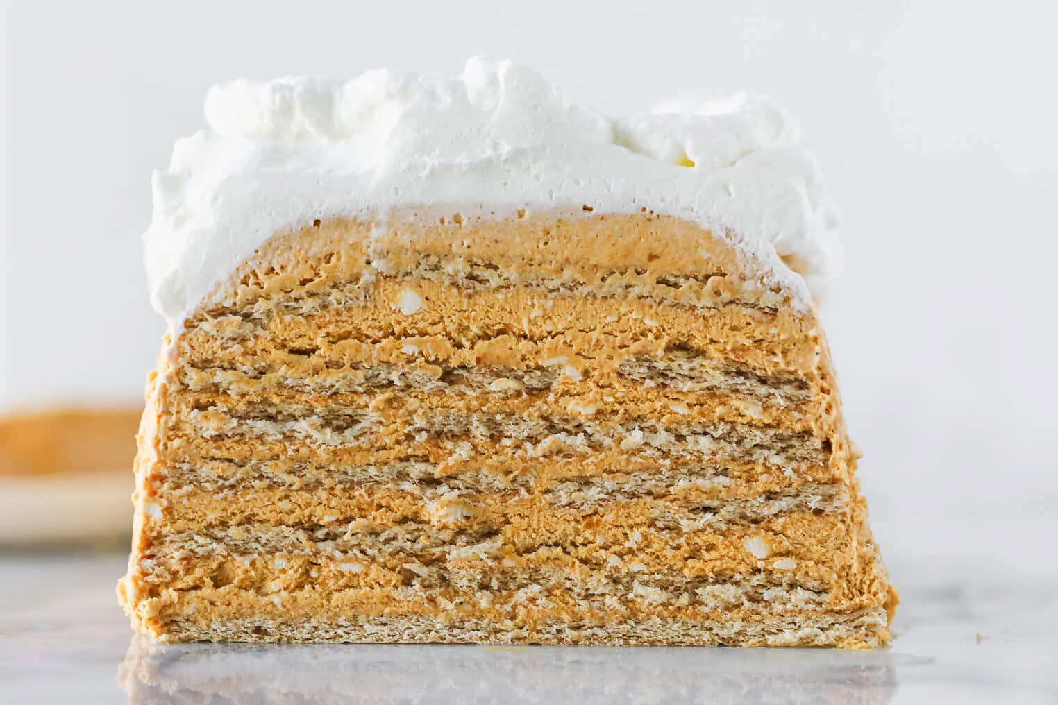 A Close-Up Of A Layered Cake With Alternating Thin, Golden-Brown Layers And Creamy Filling, Topped With A Thick Layer Of Whipped Cream, Against A Light Background.