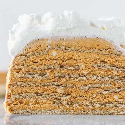 A Close-Up Of A Layered Cake With Alternating Thin, Golden-Brown Layers And Creamy Filling, Topped With A Thick Layer Of Whipped Cream, Against A Light Background.