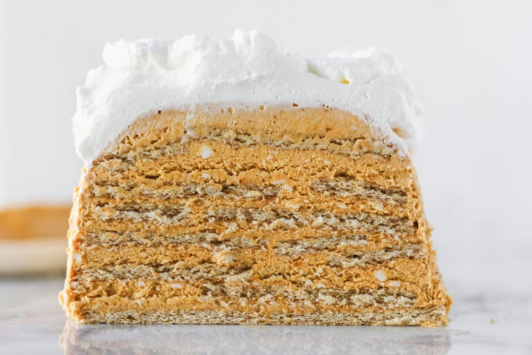 A Close-Up Of A Layered Cake With Alternating Thin, Golden-Brown Layers And Creamy Filling, Topped With A Thick Layer Of Whipped Cream, Against A Light Background.