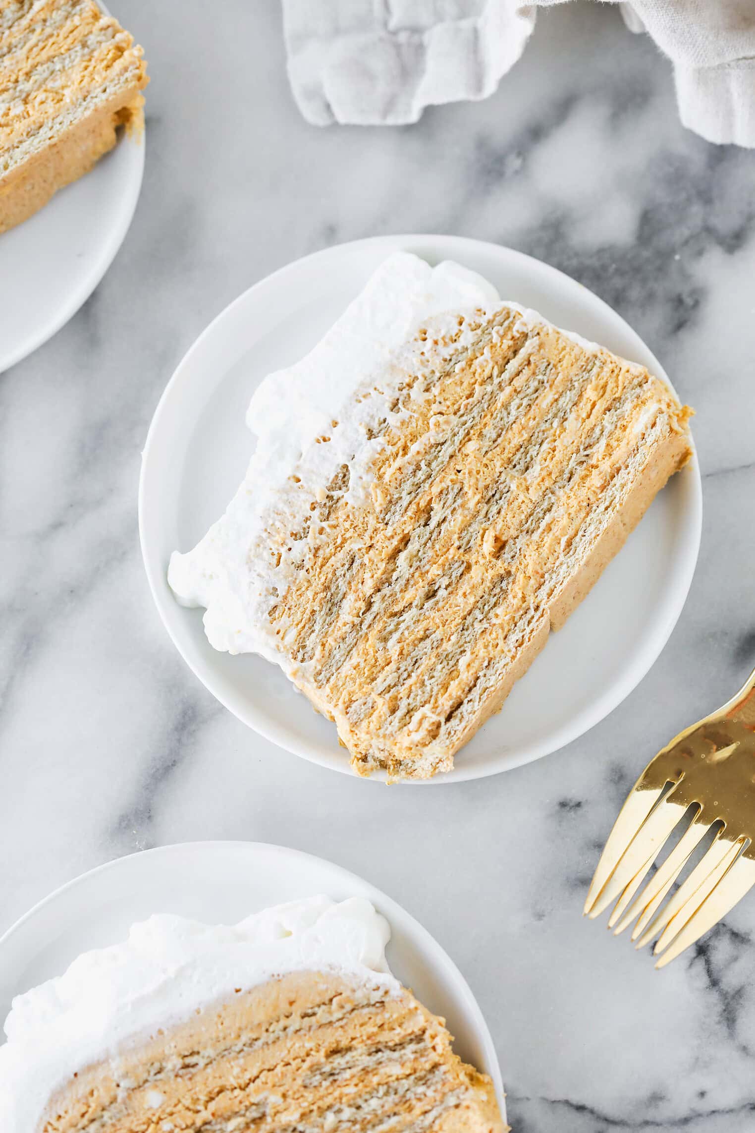 A Slice Of Layered Dessert With A Creamy Filling And A White Whipped Topping Sits On A Small White Plate, With A Gold Fork Nearby On A Marble Surface. Another Plate With Dessert Is Partially Visible.