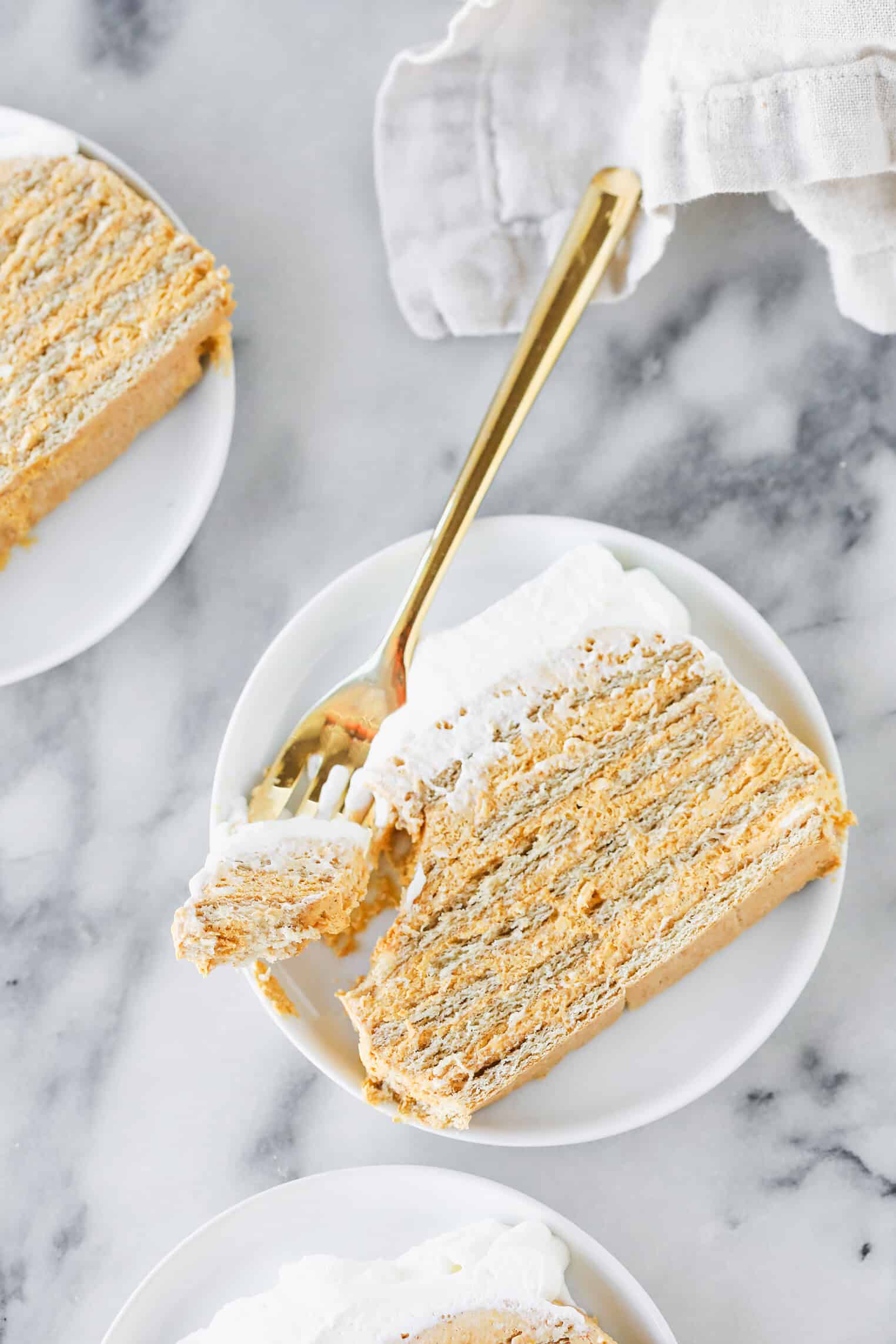 A Slice Of Layered Cake With White Frosting Sits On A White Plate, With A Gold Fork Holding A Bite. Another Cake Slice And A Folded White Napkin Are Nearby On A Marble Surface.