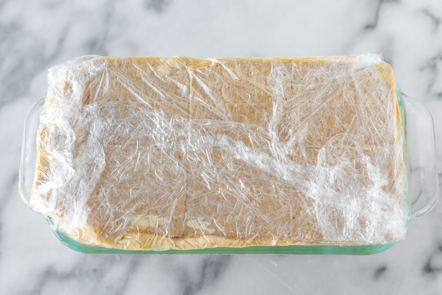 A Rectangular Glass Baking Dish Filled With A Layered Pumpkin Icebox Cake, Covered Tightly With Plastic Wrap, Sits On A Marble Surface.