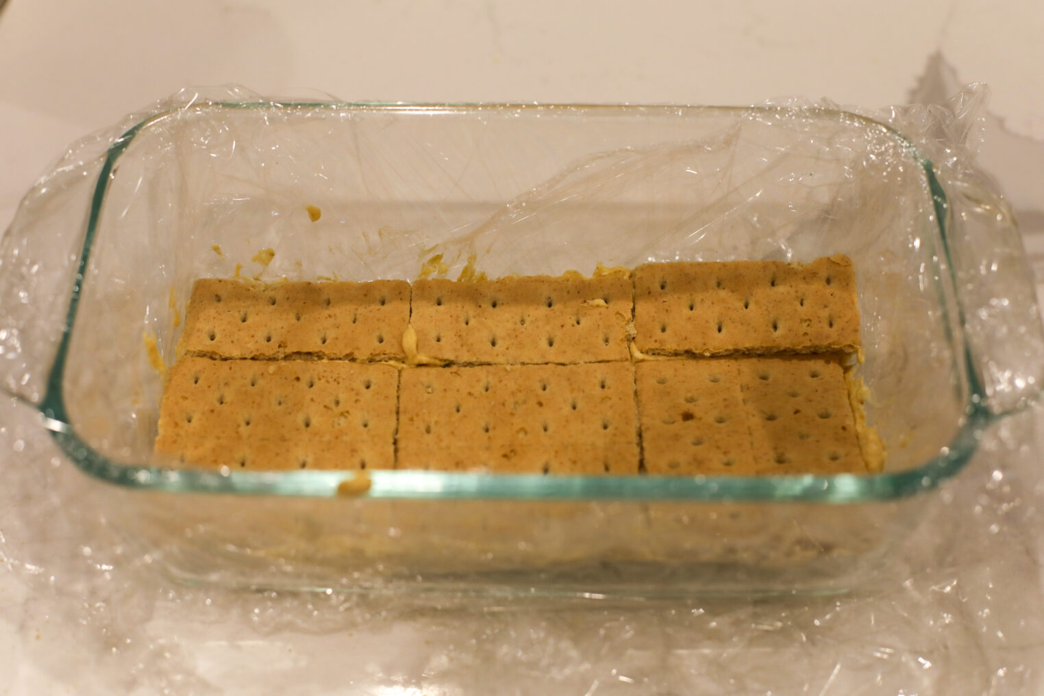A Glass Dish Lined With Plastic Wrap Contains A Single Layer Of Rectangular Graham Crackers, Perfect As The Base For A Delicious Pumpkin Icebox Cake.