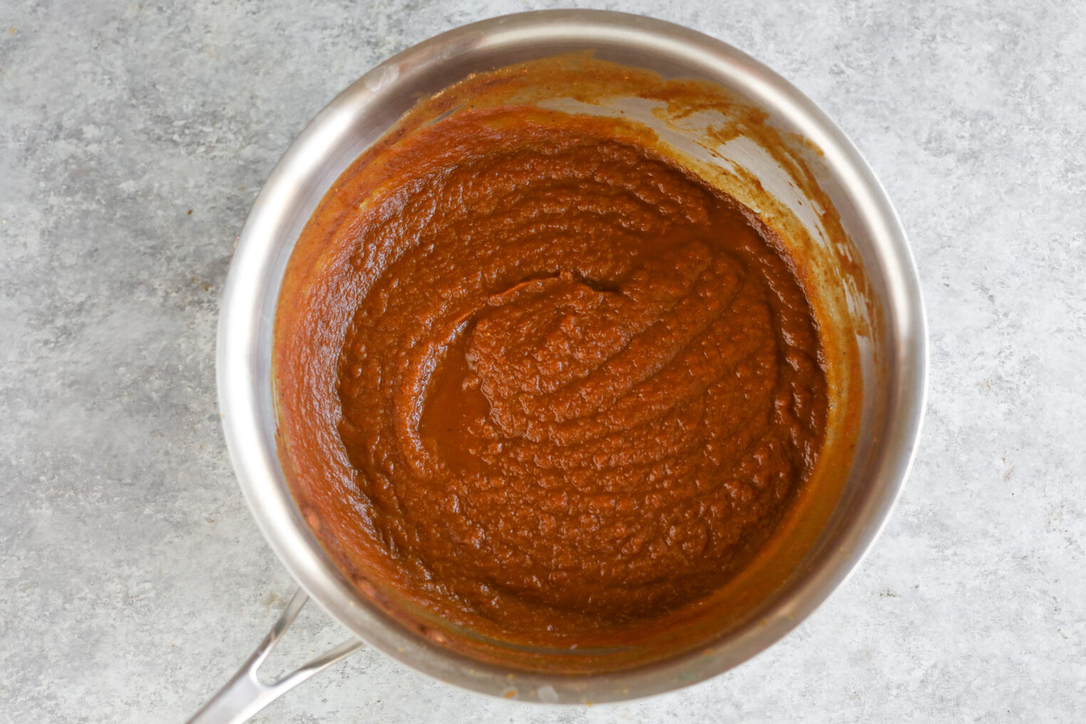 A Stainless Steel Saucepan Filled With Thick, Reddish-Brown Sauce Or Paste For Pumpkin Icebox Cake Sits On A Gray Textured Surface. The Sauce Has A Smooth, Slightly Chunky Texture And Is Swirled In The Pan.