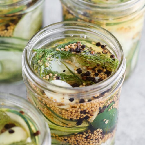 Glass Jars Filled With Sliced Cucumbers, Garlic Cloves, Dill, Mustard Seeds, And Black Peppercorns Sit On A Light Gray Surface—Perfect For Making Delicious Refrigerator Pickles.