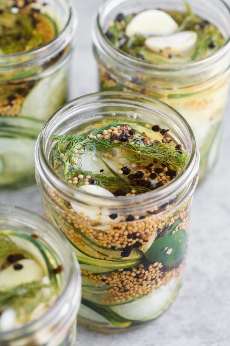 Glass Jars Filled With Sliced Cucumbers, Garlic Cloves, Dill, Mustard Seeds, And Black Peppercorns Sit On A Light Gray Surface—Perfect For Making Delicious Refrigerator Pickles.