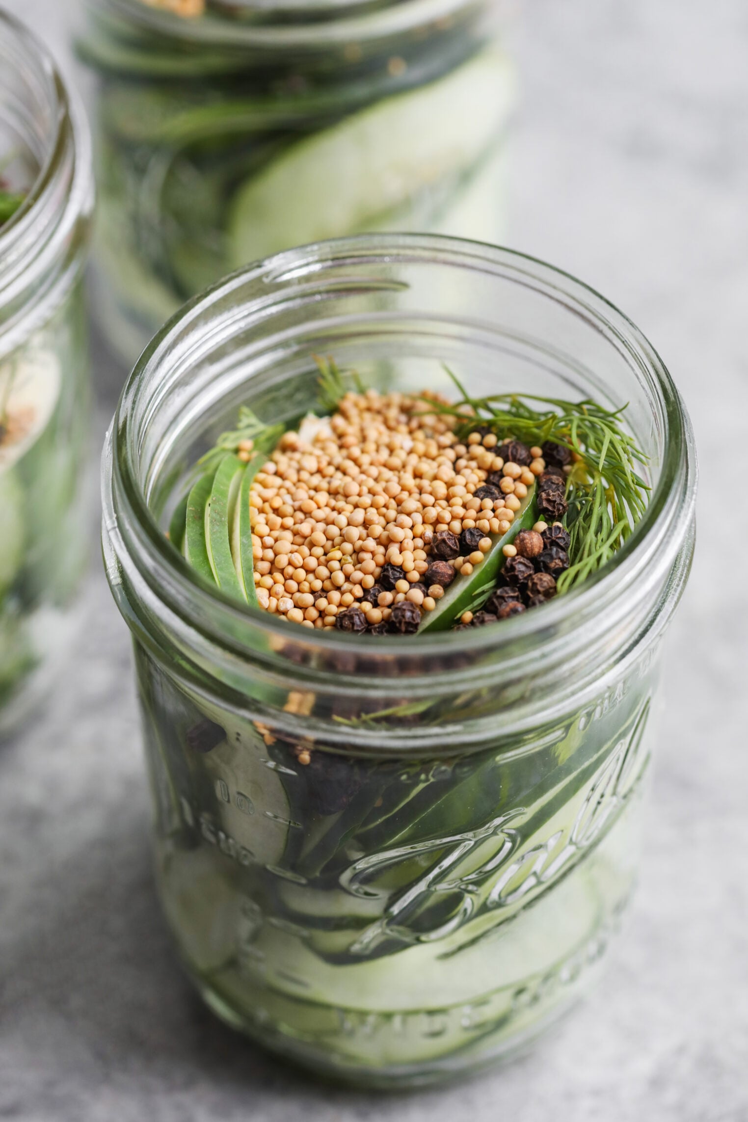 A Glass Mason Jar Filled With Cucumber Slices, Fresh Dill, Mustard Seeds, Black Peppercorns, And Other Pickling Spices—Perfect For Making Refrigerator Pickles. Another Jar Is Partially Visible In The Background.