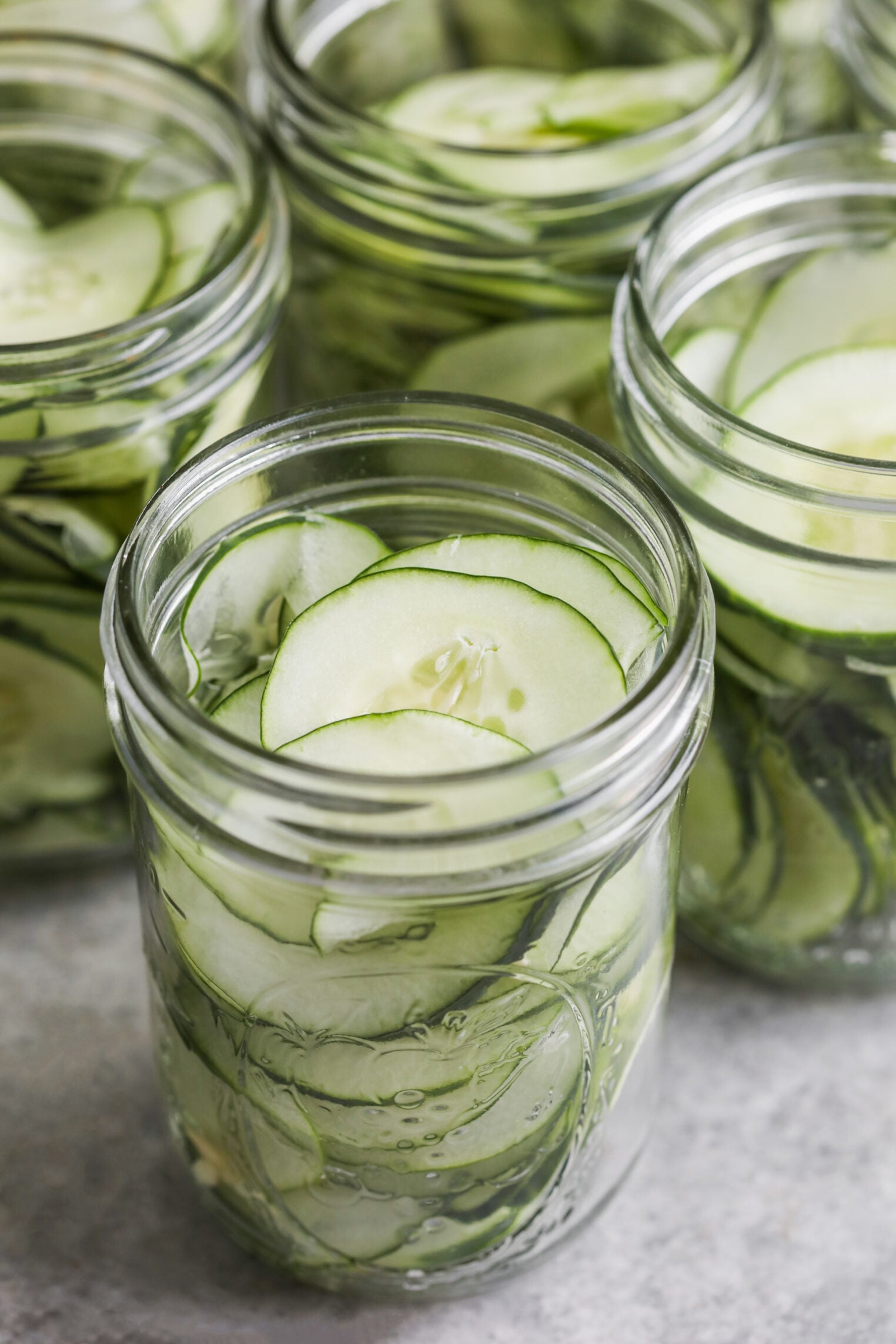 Several Glass Jars Filled With Thinly Sliced Cucumbers Stacked Vertically, Ready To Become Refrigerator Pickles, Sit On A Gray Surface.