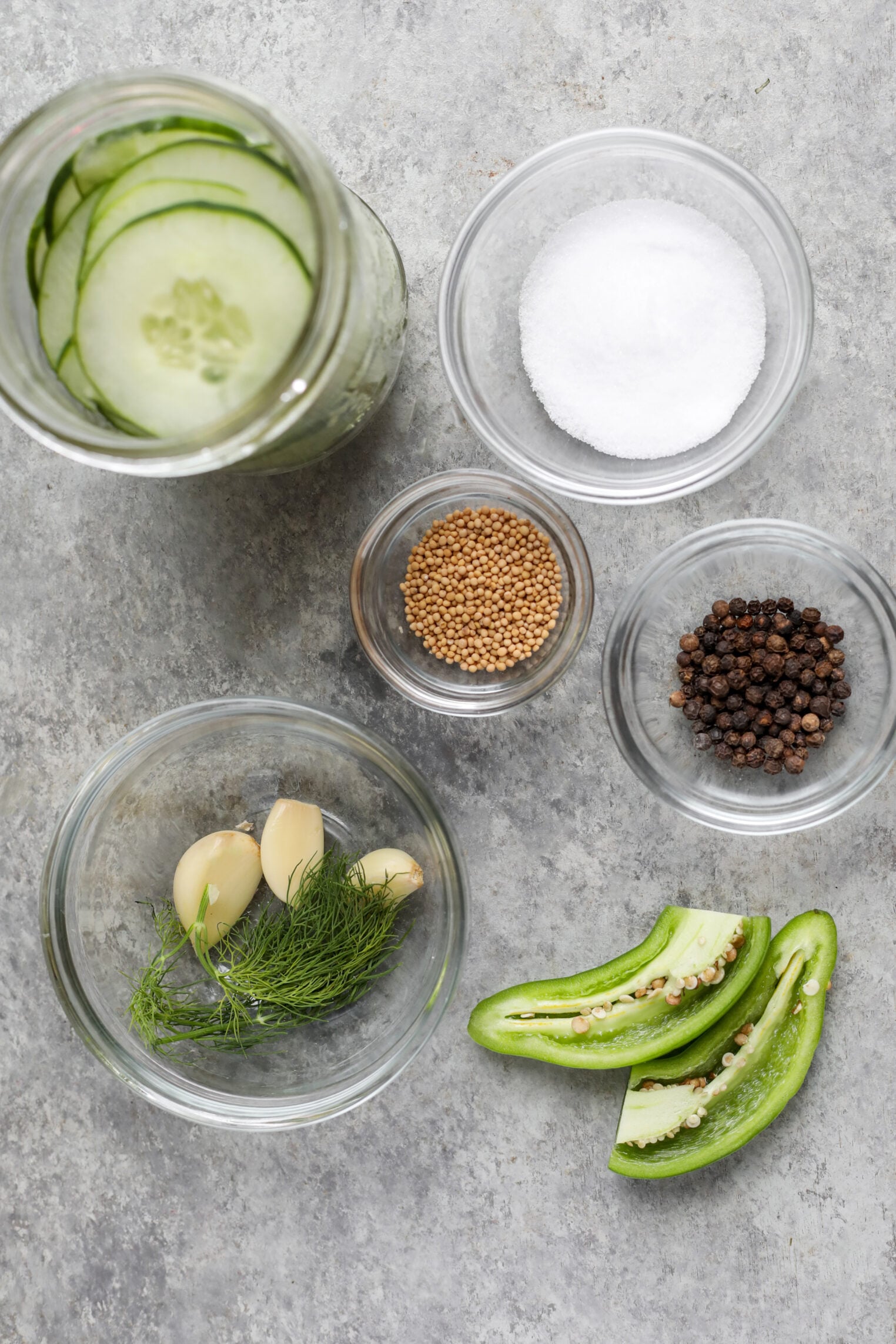 Several Clear Bowls On A Gray Surface Contain Ingredients For Refrigerator Pickles: Cucumber Slices, Salt, Mustard Seeds, Black Peppercorns, Garlic Cloves With Dill, And Sliced Green Peppers.