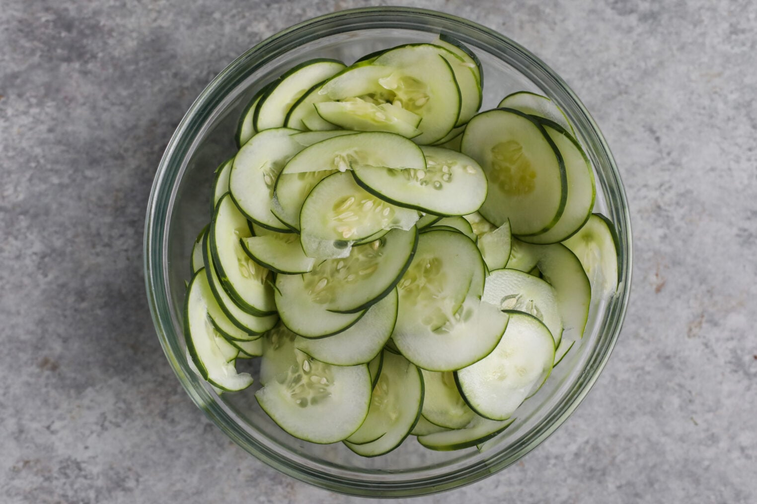 A Glass Bowl Filled With Thinly Sliced Cucumbers For Refrigerator Pickles, Viewed From Above, Sits On A Gray Textured Surface.