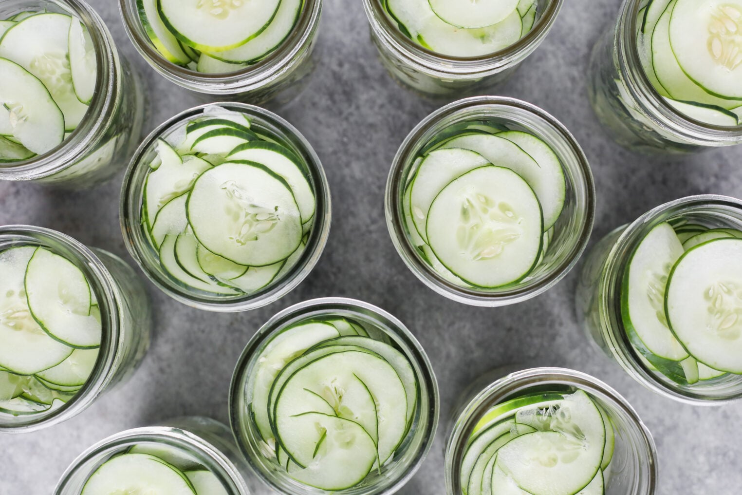 Several Glass Jars Filled With Thinly Sliced Cucumbers, Perfect For Making Refrigerator Pickles, Are Viewed From Above And Arranged Closely Together On A Gray Surface.