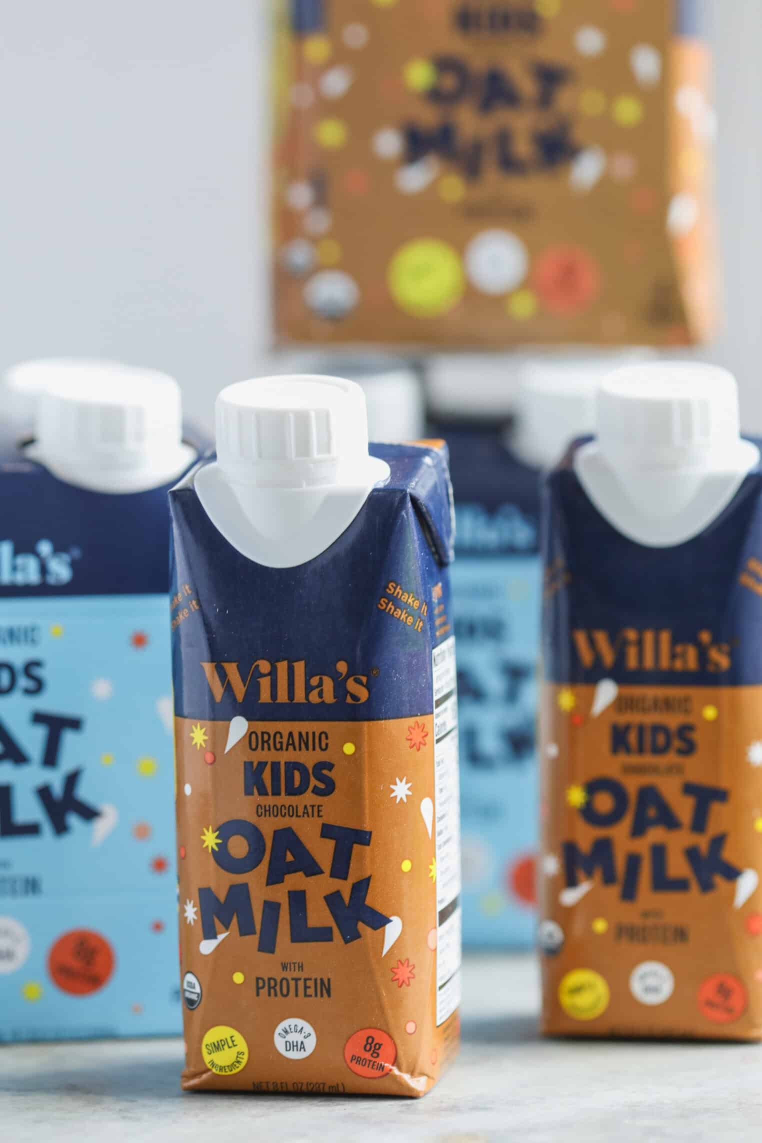 Registered Dietitian Approved Road Trip Summer Snacks 1 Three Cartons Of Willa’s Organic Kids Oat Milk Are In Focus, With A Chocolate Flavor Carton In Front And Two Blue Original Cartons Behind—Perfect As Summer Road Trip Snacks. Blurred Oat Milk Cartons Sit In The Background.