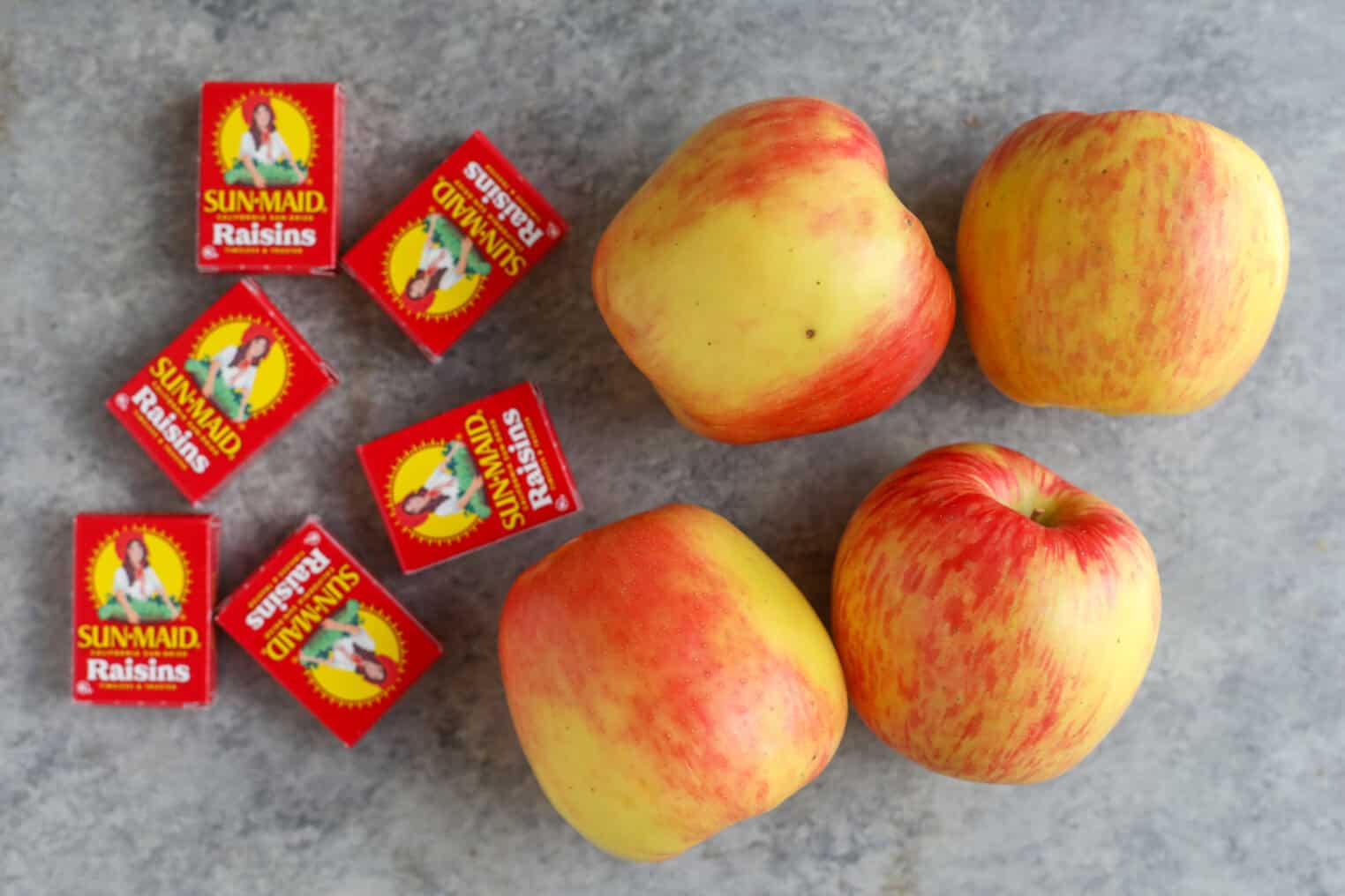 Registered Dietitian Approved Road Trip Summer Snacks 3 Four Red And Yellow Apples And Six Small Red Boxes Of Sun-Maid Raisins, Perfect As Summer Road Trip Snacks, Are Arranged On A Gray Surface.