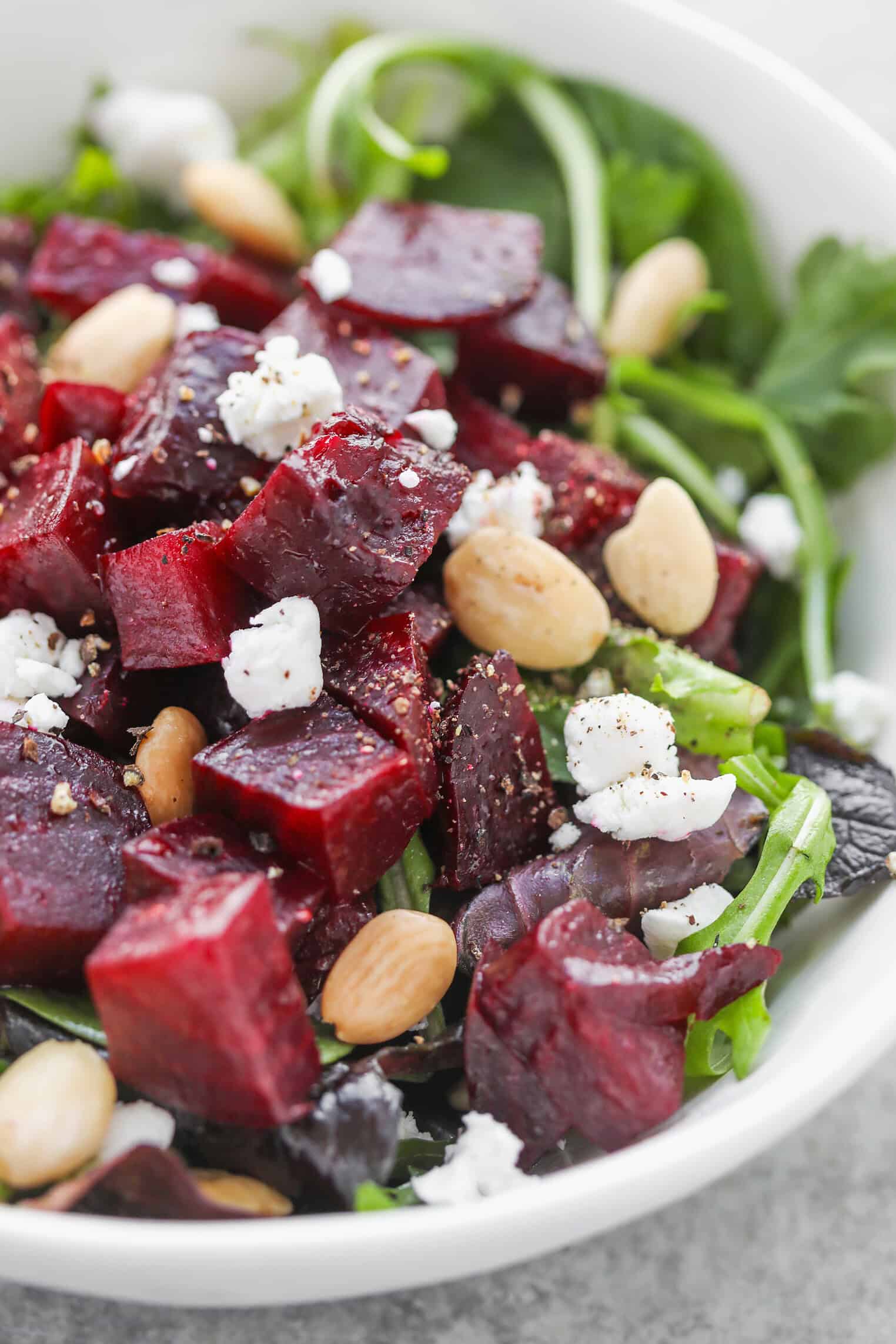 A Roasted Beet Salad With Diced Red Beets, Almonds, And Crumbled Feta Cheese Sits On A Bed Of Mixed Greens In A White Bowl. Freshly Ground Pepper Is Sprinkled On Top.